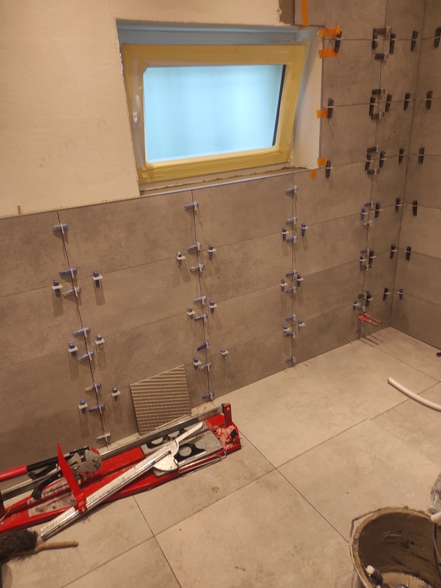 Układanie szarych płytek na ścianie w łazience z oknem. Widoczne krzyżyki dystansowe, poziomica i wiadro z zaprawą. Praca w toku, etap kładzenia glazury.