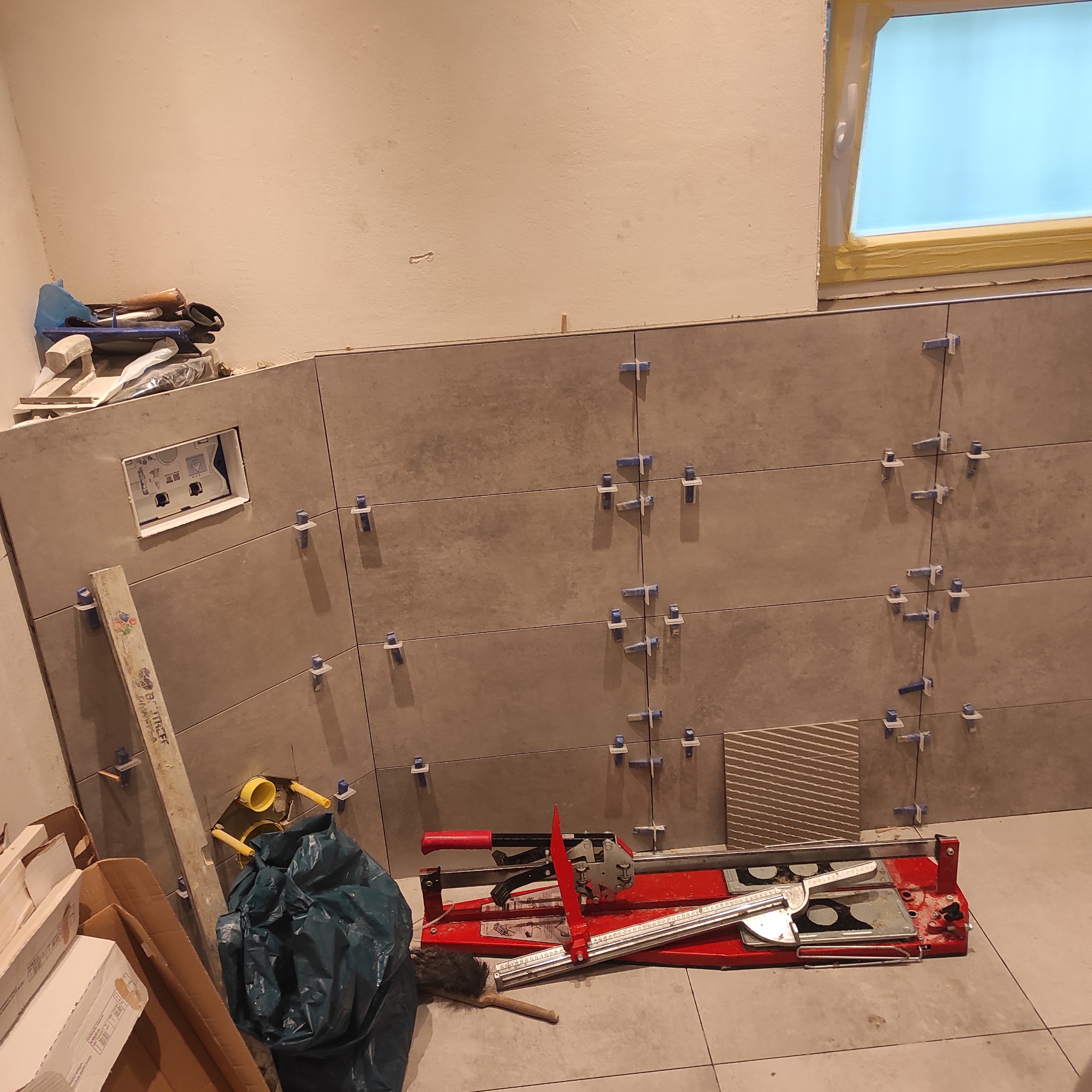 Montaż płytek ściennych w łazience, widoczne narzędzia glazurnicze, poziomica,  krzyżyki dystansowe i  fragment okna zabezpieczonego taśmą malarską.