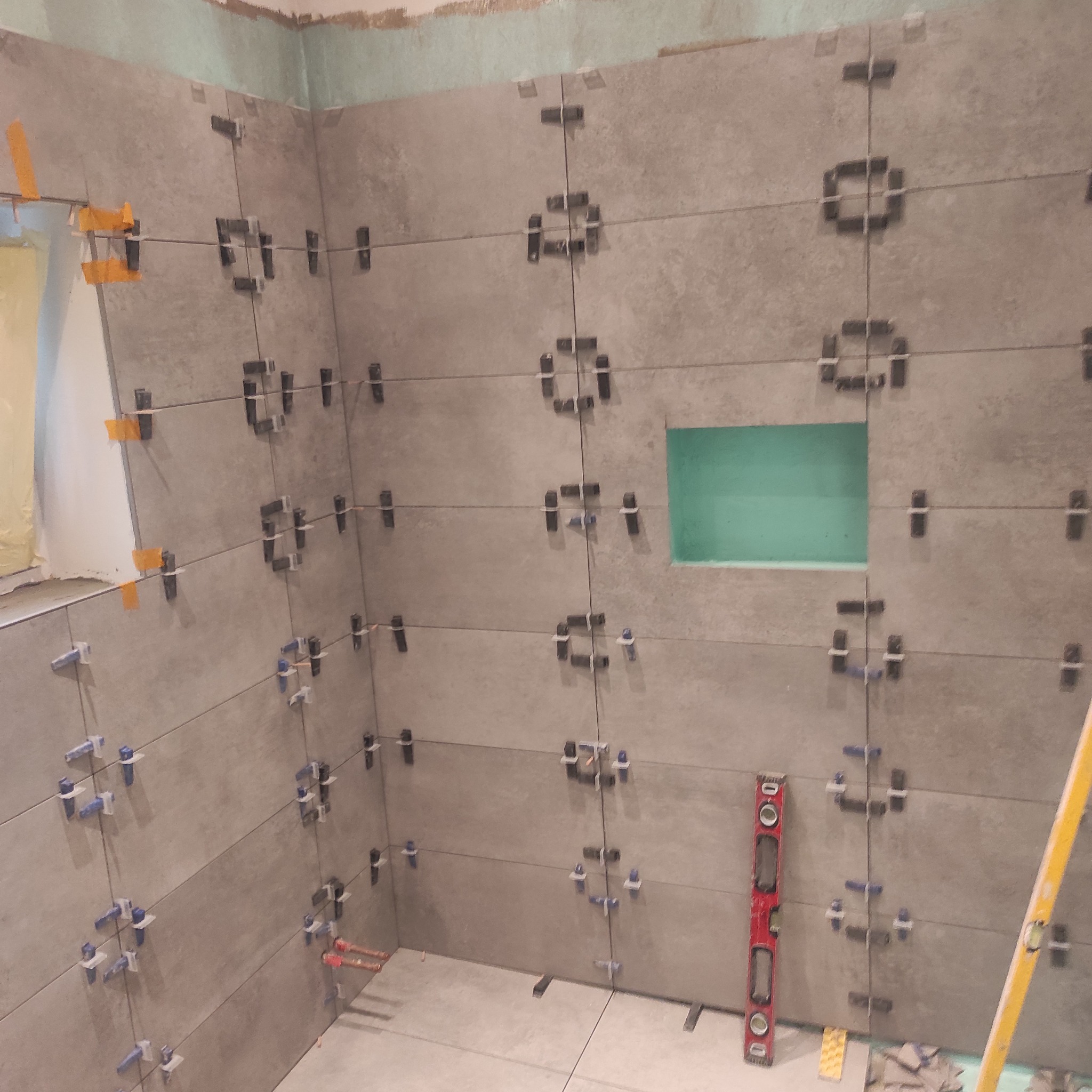 Ściana łazienki w trakcie wykańczania z szarymi płytkami, krzyżykami dystansowymi i poziomicą. Widoczny fragment wnęki. Etap prac przed fugowaniem.