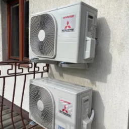 Dwa klimatyzatory Mitsubishi Inverter zamontowane na elewacji budynku, widoczne z zewnątrz, z balkonem z metalową balustradą i oknem w tle.
