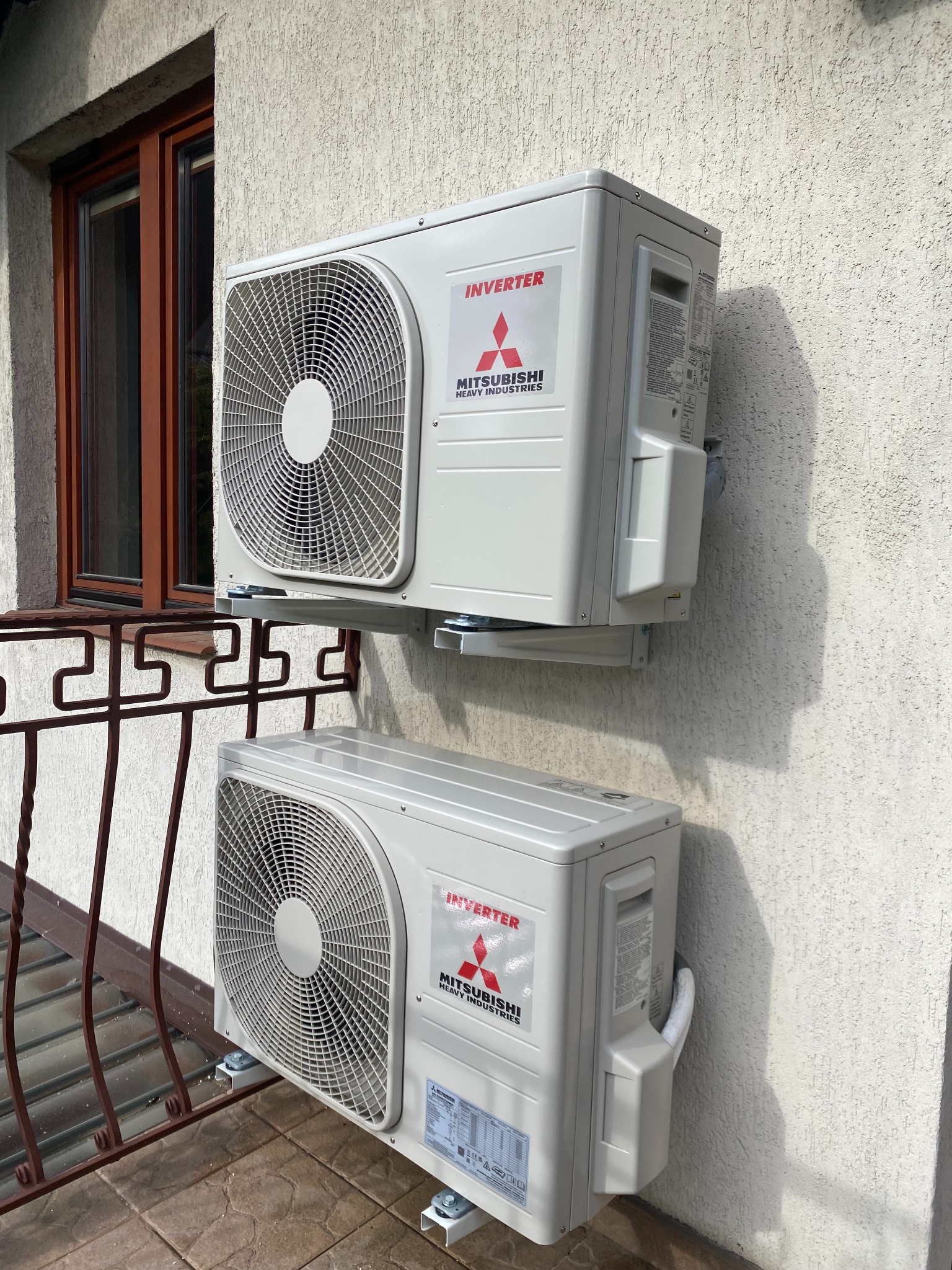 Dwa klimatyzatory Mitsubishi Inverter zamontowane na elewacji budynku, widoczne z zewnątrz, z balkonem z metalową balustradą i oknem w tle.