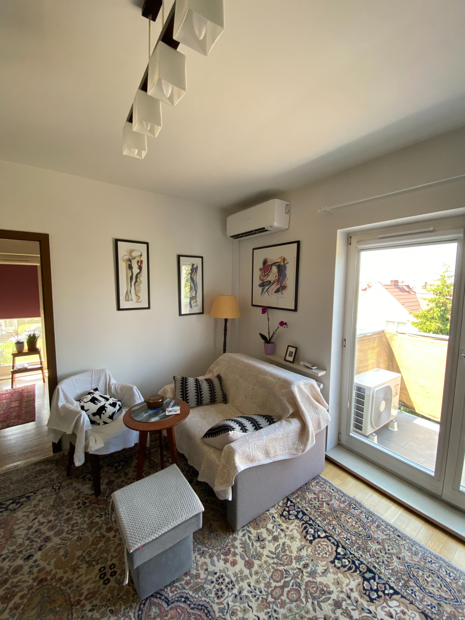 Klimatyzator ścienny w jasnym salonie z wyjściem na balkon. Widoczny również klimatyzator na balkonie. Wnętrze urządzone w stylu retro.