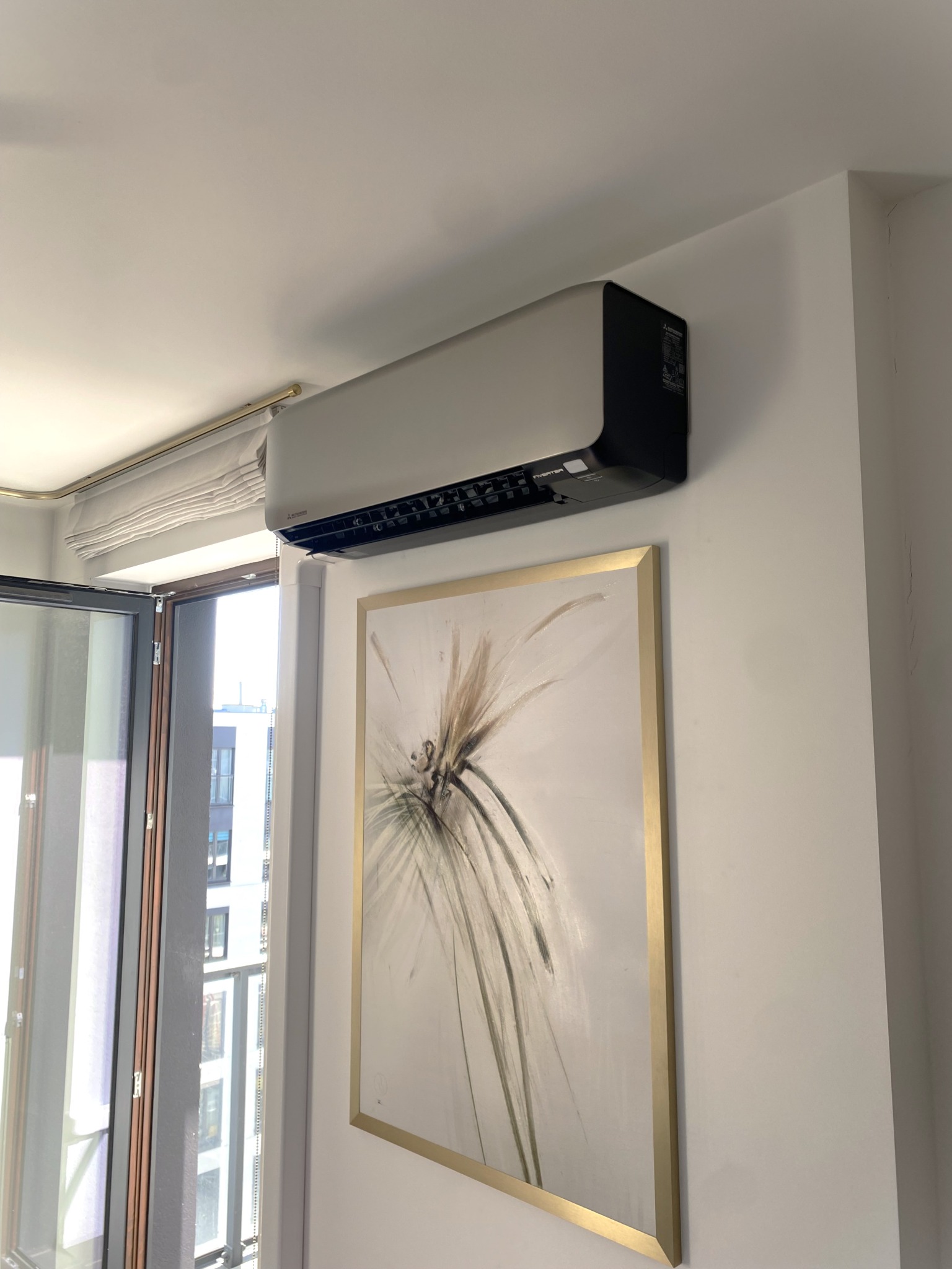 Szara klimatyzacja Mitsubishi na ścianie nad obrazem w złotej ramie, widok z boku, okno z roletą w tle, jasne wnętrze.