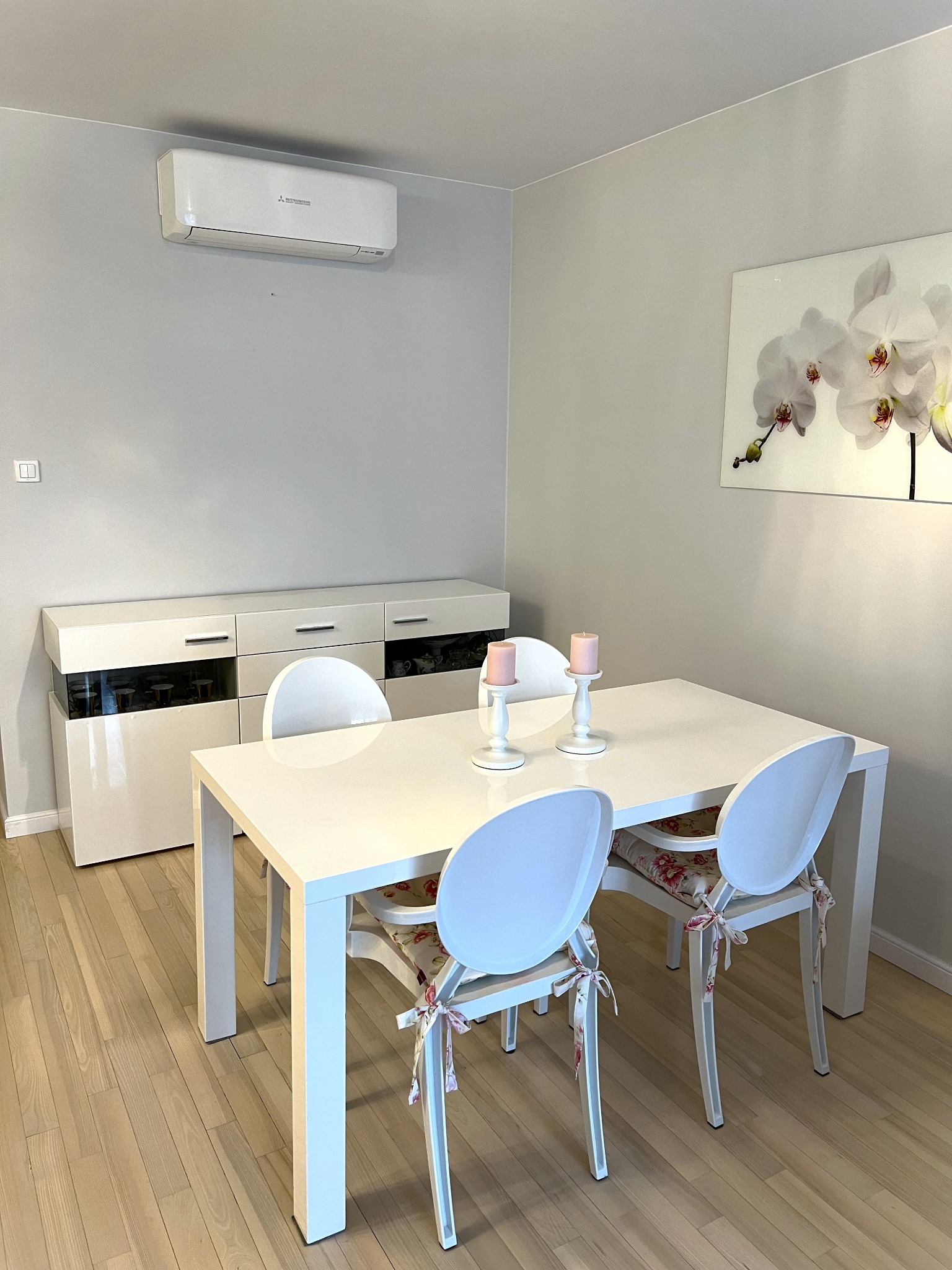 Biały klimatyzator Mitsubishi na ścianie nad białym kredensem w jadalni z białym stołem i krzesłami. Na stole świeczki. Obraz z motywem kwiatowym na ścianie.