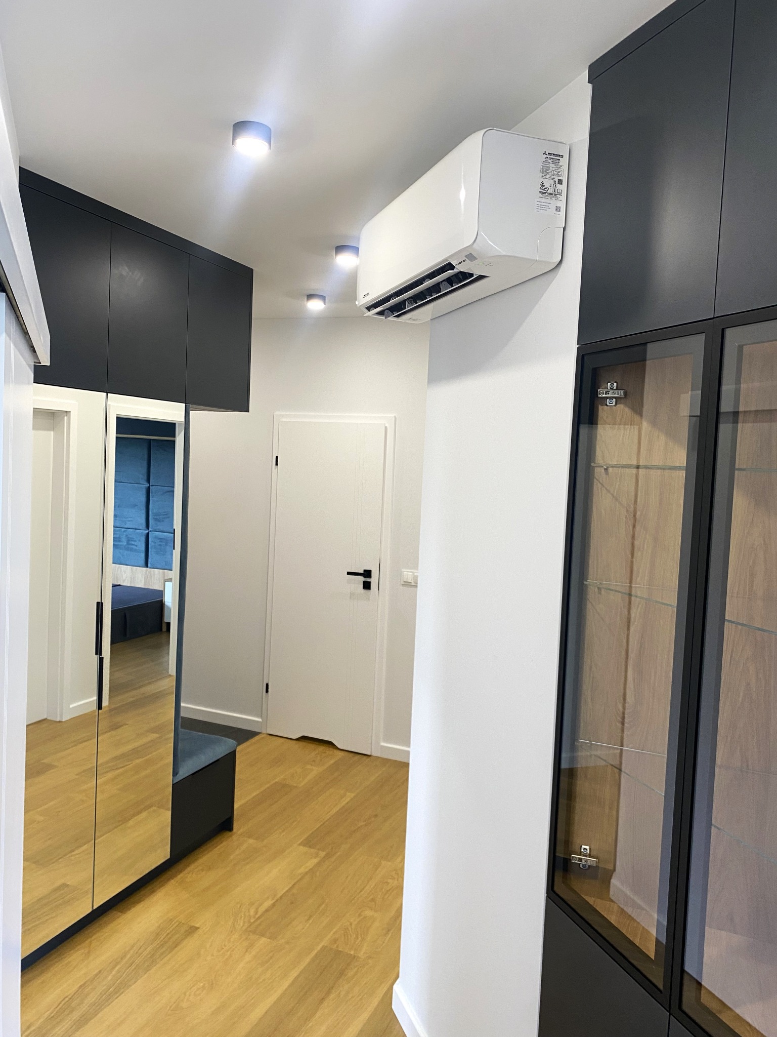Biała klimatyzacja ścienna w nowoczesnym przedpokoju z szarą zabudową i lustrem. Widoczne oświetlenie sufitowe i drewniana podłoga.