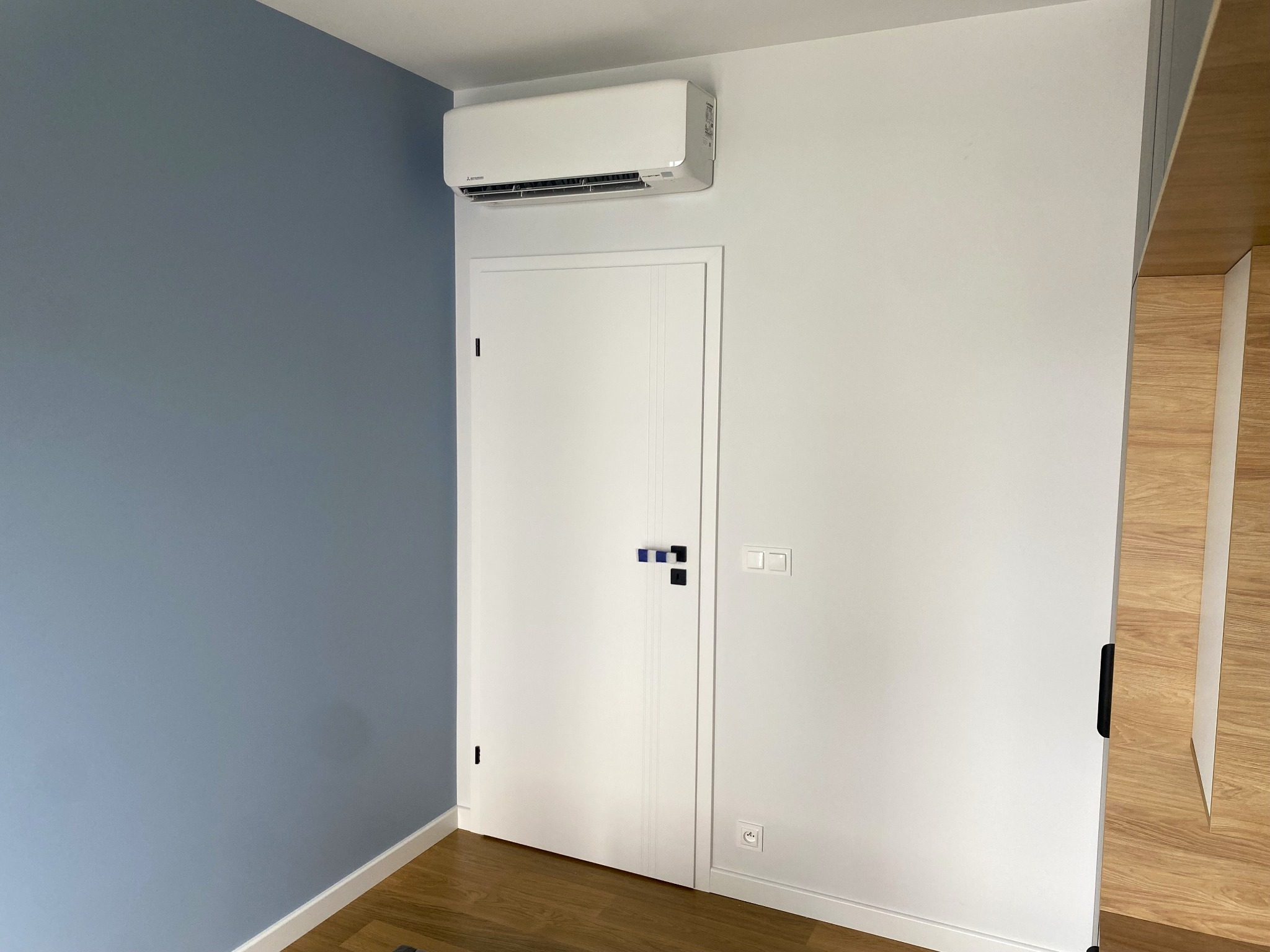 Nowoczesny klimatyzator Mitsubishi zainstalowany nad białymi drzwiami w pomieszczeniu z błękitną ścianą. Widoczne gniazdka i włącznik światła.