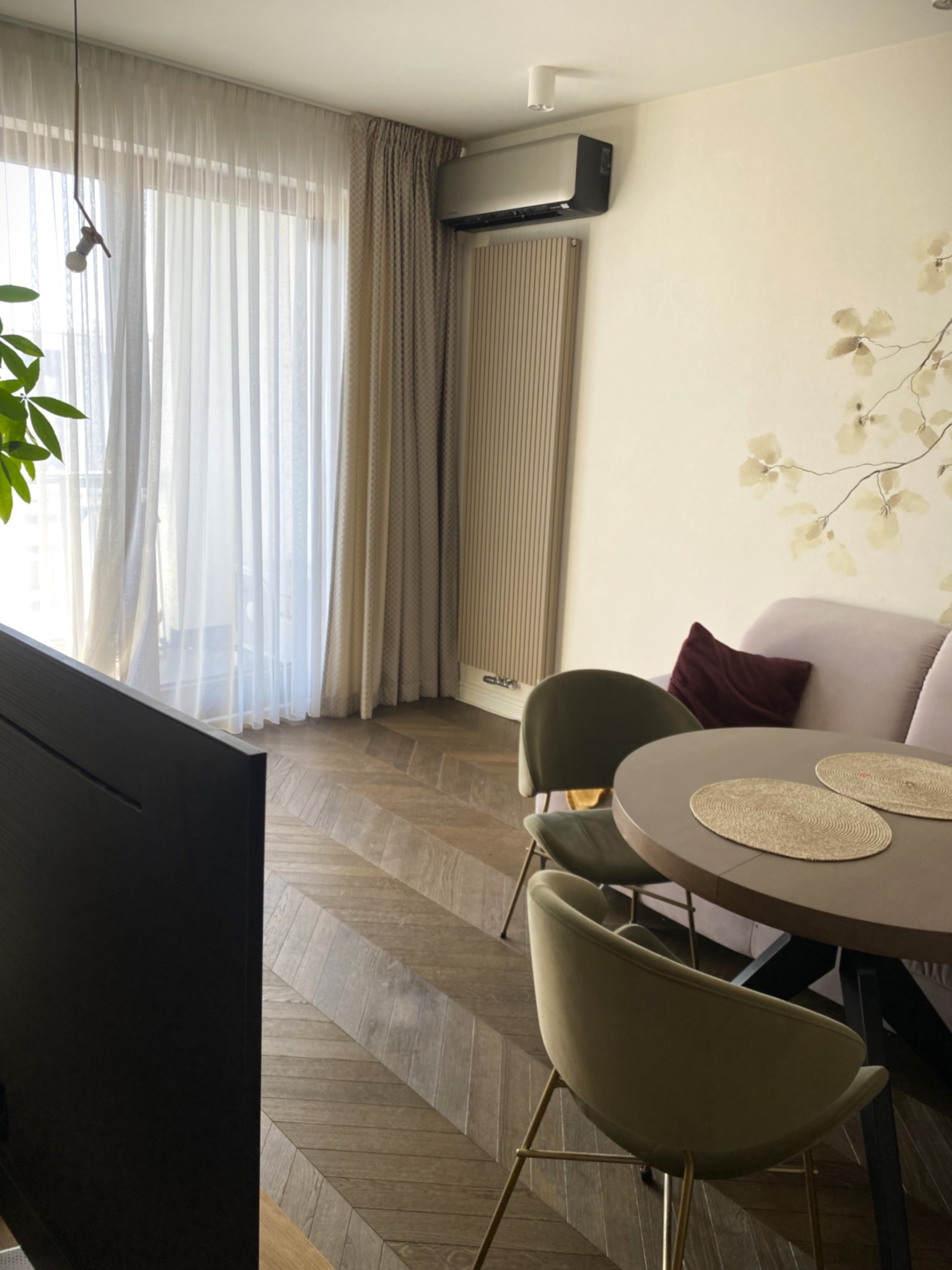 Eleganckie wnętrze: klimatyzator ścienny nad grzejnikiem, okno z firanami, stół z krzesłami, parkiet w jodełkę i dekoracja kwiatowa na ścianie.