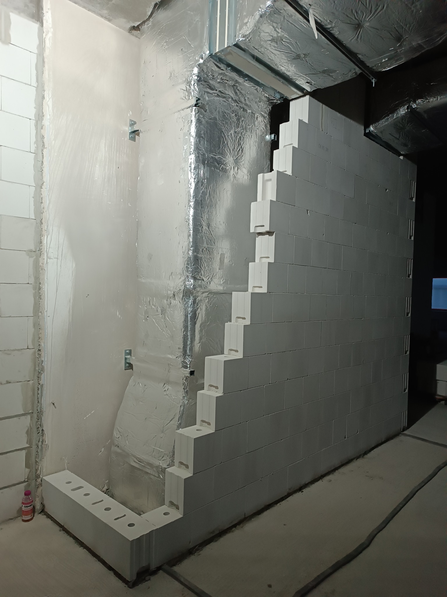 Wznoszenie ściany z białych bloczków gazobetonowych w surowym wnętrzu. Widoczne fragmenty instalacji i izolacji. Ściana w trakcie budowy, z widocznymi stopniami.