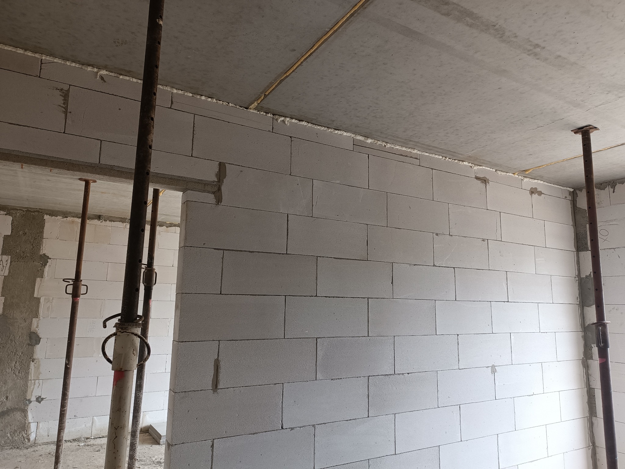 Wnętrze w trakcie budowy: ściana z białych bloczków, podpory stropowe, surowy strop. Widoczny etap murowania ścian z widocznymi spoinami i pianką montażową.