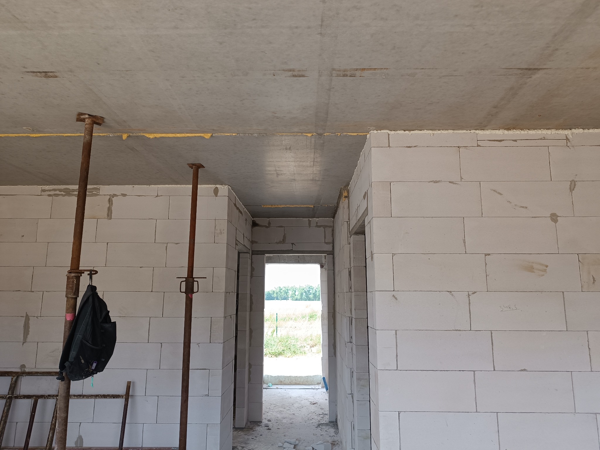 Wnętrze w trakcie budowy: ściany z betonu komórkowego, stalowe podpory, widok na zewnątrz przez otwory drzwiowe. Surowy stan, w tle zieleń.