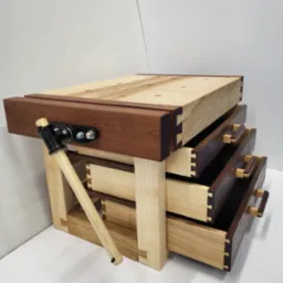 Drewniany stół warsztatowy z trzema szufladami, widoczne połączenia na jaskółczy ogon, z imadłem i młotkiem, wykonany z różnych gatunków drewna.