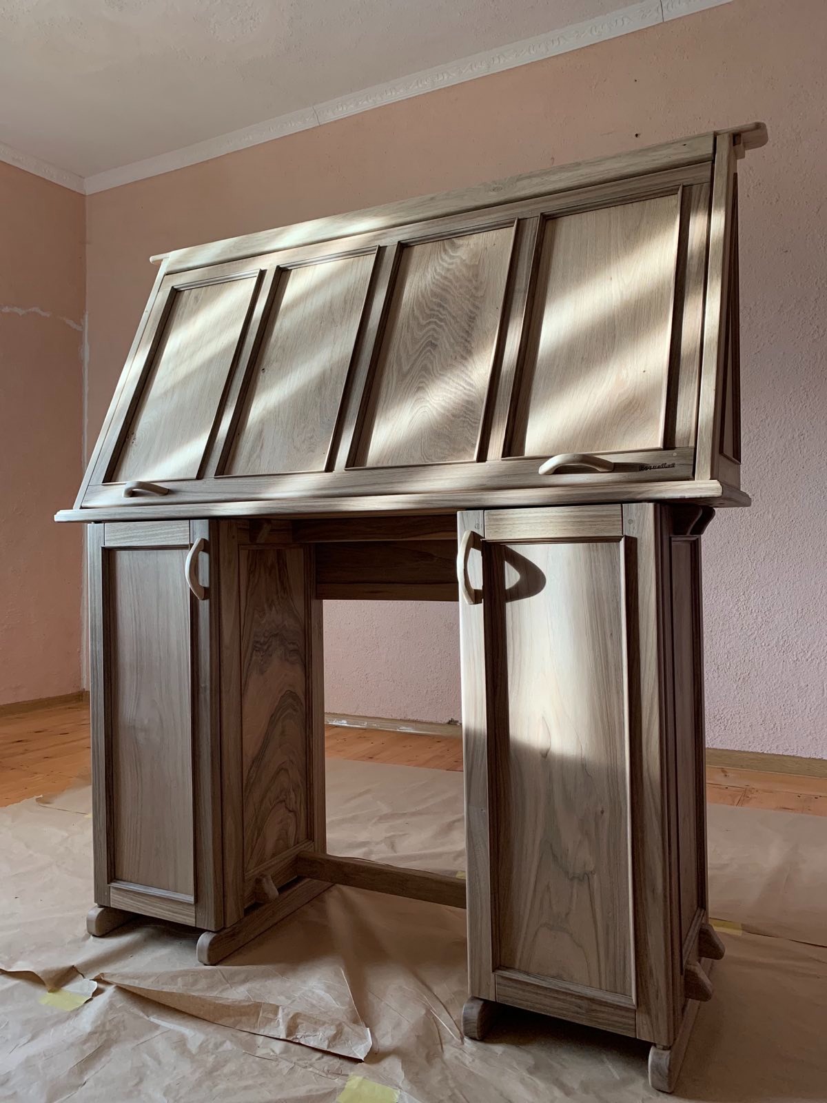 Drewniane biurko sekretarzyk z szufladami i klapą, oświetlone promieniami słońca, stoi na papierze ochronnym w pokoju z różową ścianą.