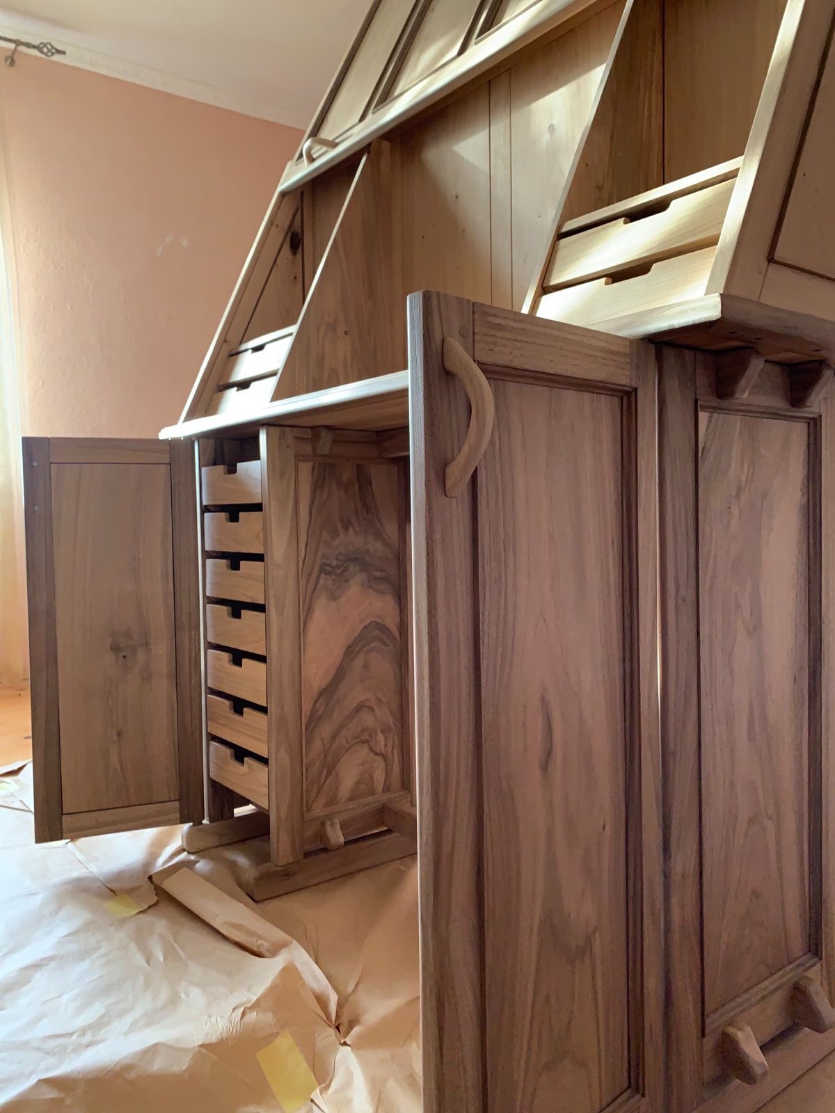 Drewniany sekretarzyk z otwartymi drzwiczkami i szufladami, widoczne detale wykończenia, zabezpieczony papierem malarskim, ciepłe oświetlenie.