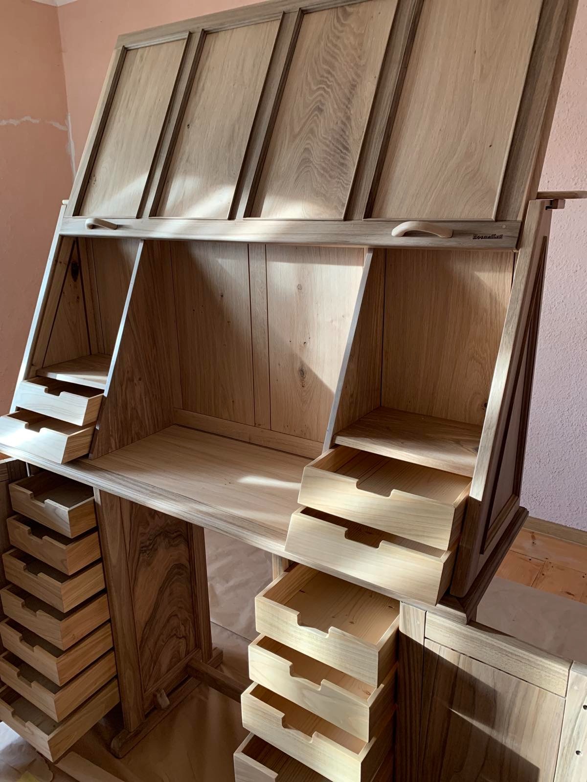 Drewniane biurko-sekretarzyk z licznymi szufladami, wykonane z jasnego drewna. Górna część biurka jest podnoszona, tworząc pochyloną powierzchnię do pisania.