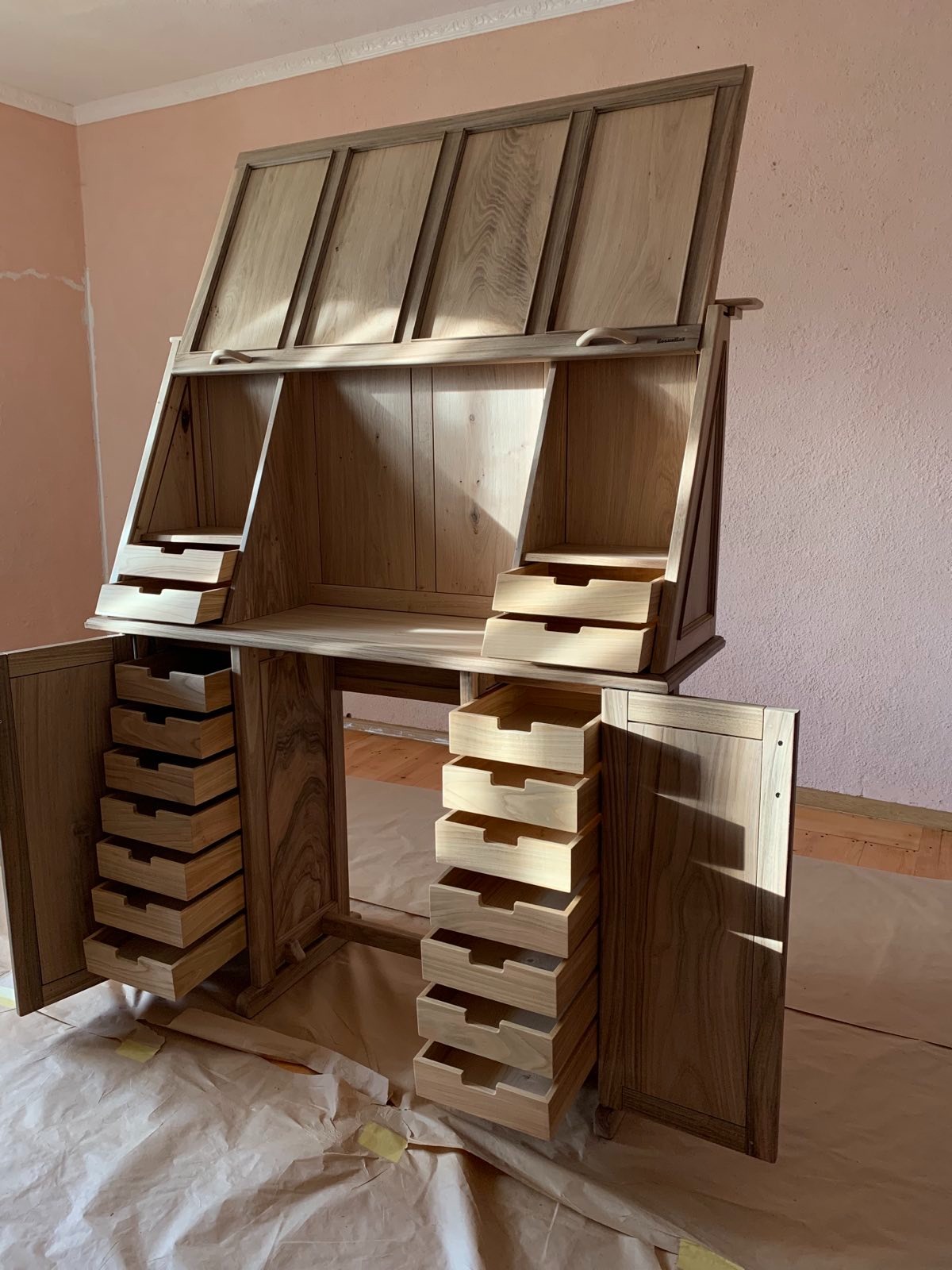 Drewniane biurko sekretarzyk z otwartymi drzwiczkami i szufladami, widoczne usłojenie drewna, zabezpieczone podłoże, surowe wykończenie.