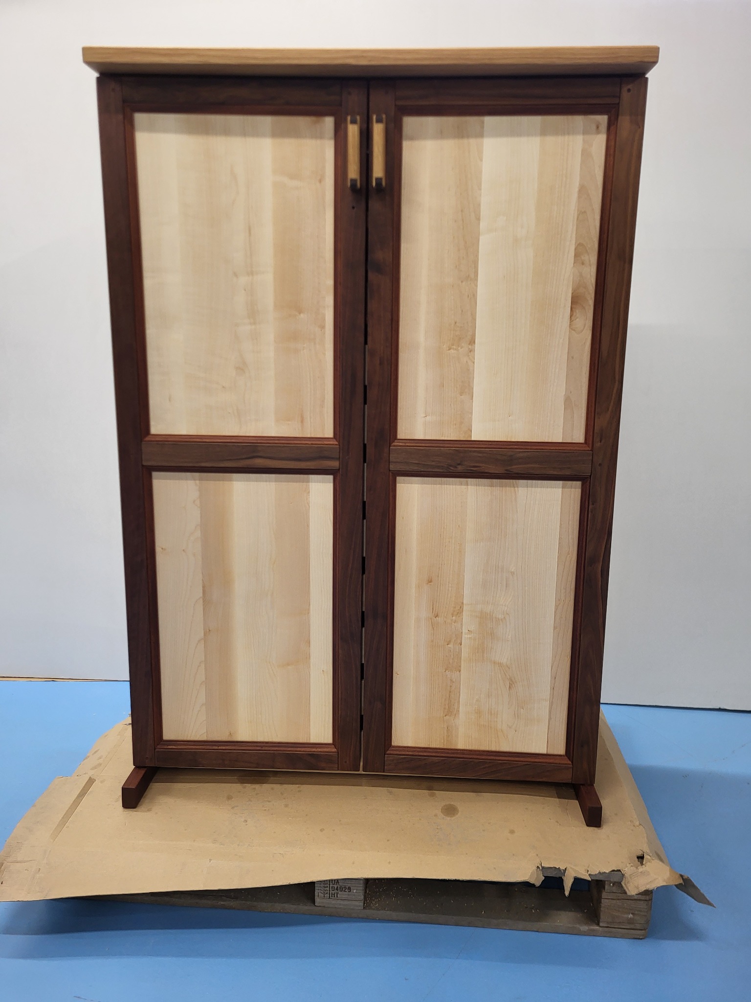 Dwudrzwiowa szafka z jasnego drewna z ciemnobrązowymi ramami, stoi na palecie. Minimalistyczny design, widoczne usłojenie drewna, proste uchwyty.