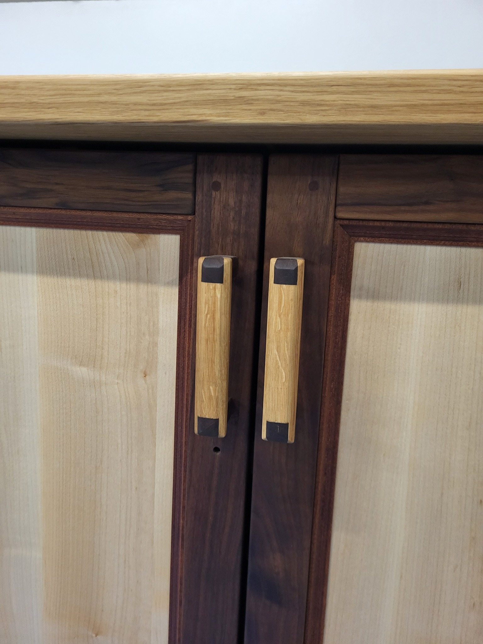Dwudrzwiowa szafka z jasnego drewna i ciemnymi ramami, z unikalnymi, pionowymi uchwytami z jasnego drewna z ciemnymi akcentami na końcach.