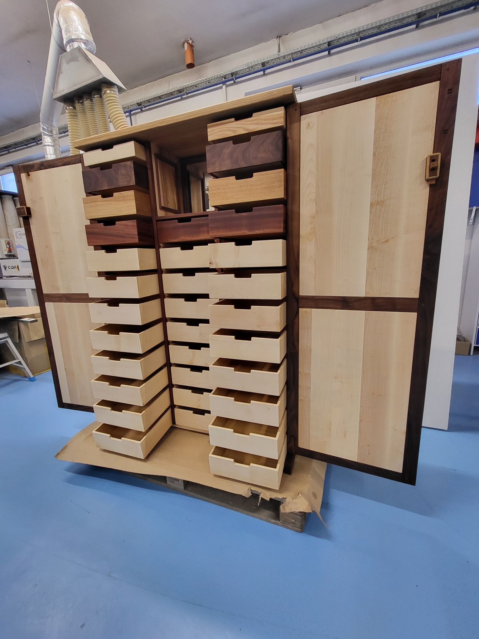 Otwarta szafa z jasnego drewna z ciemnymi obramowaniami i rzędami szuflad w dwóch odcieniach brązu, stojąca na palecie w warsztacie.