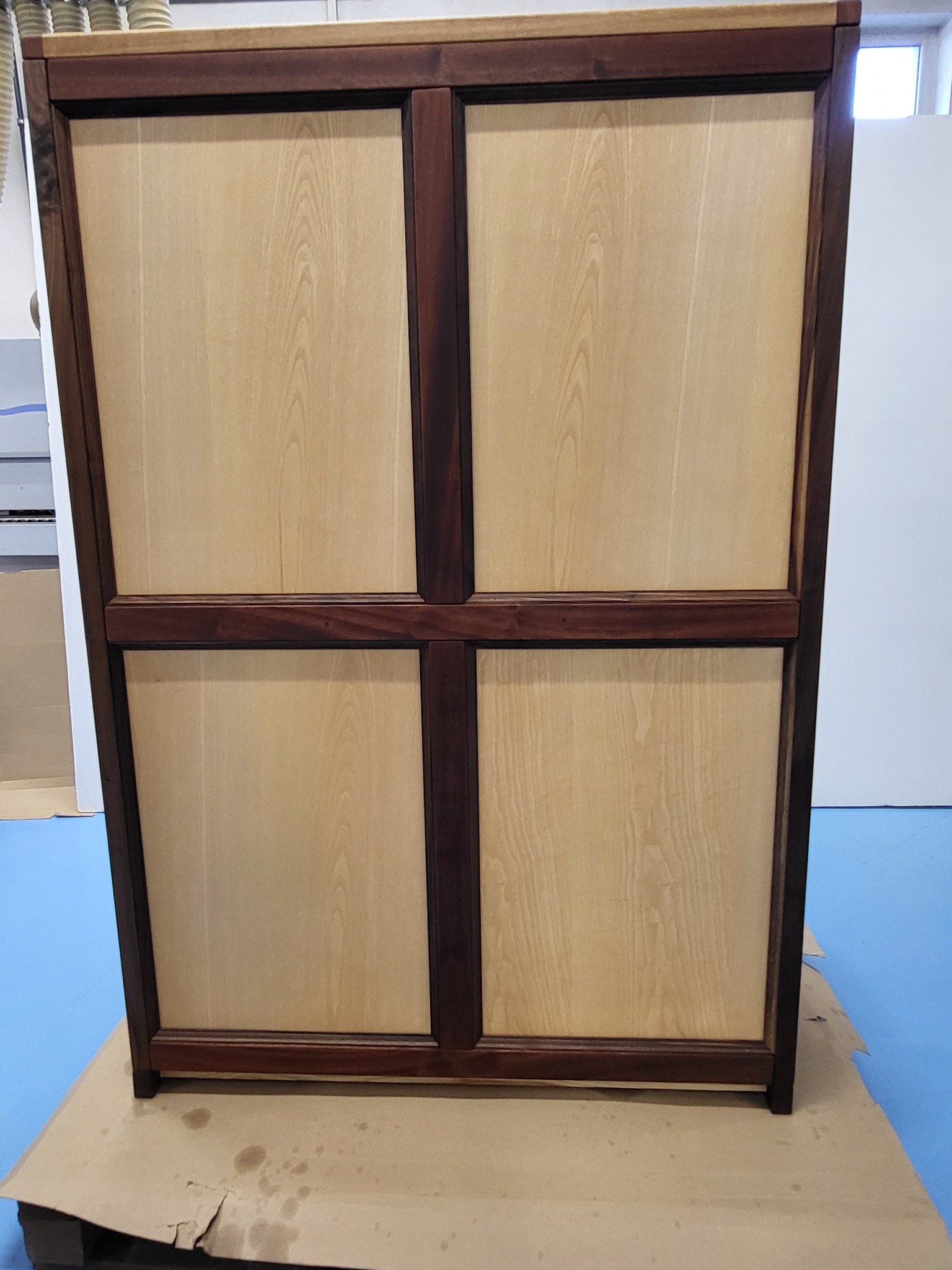 Dwudrzwiowa szafka z jasnego drewna z ciemnobrązowymi ramami, stoi na tekturze na niebieskiej podłodze. Widoczne słoje drewna i detale wykończenia.