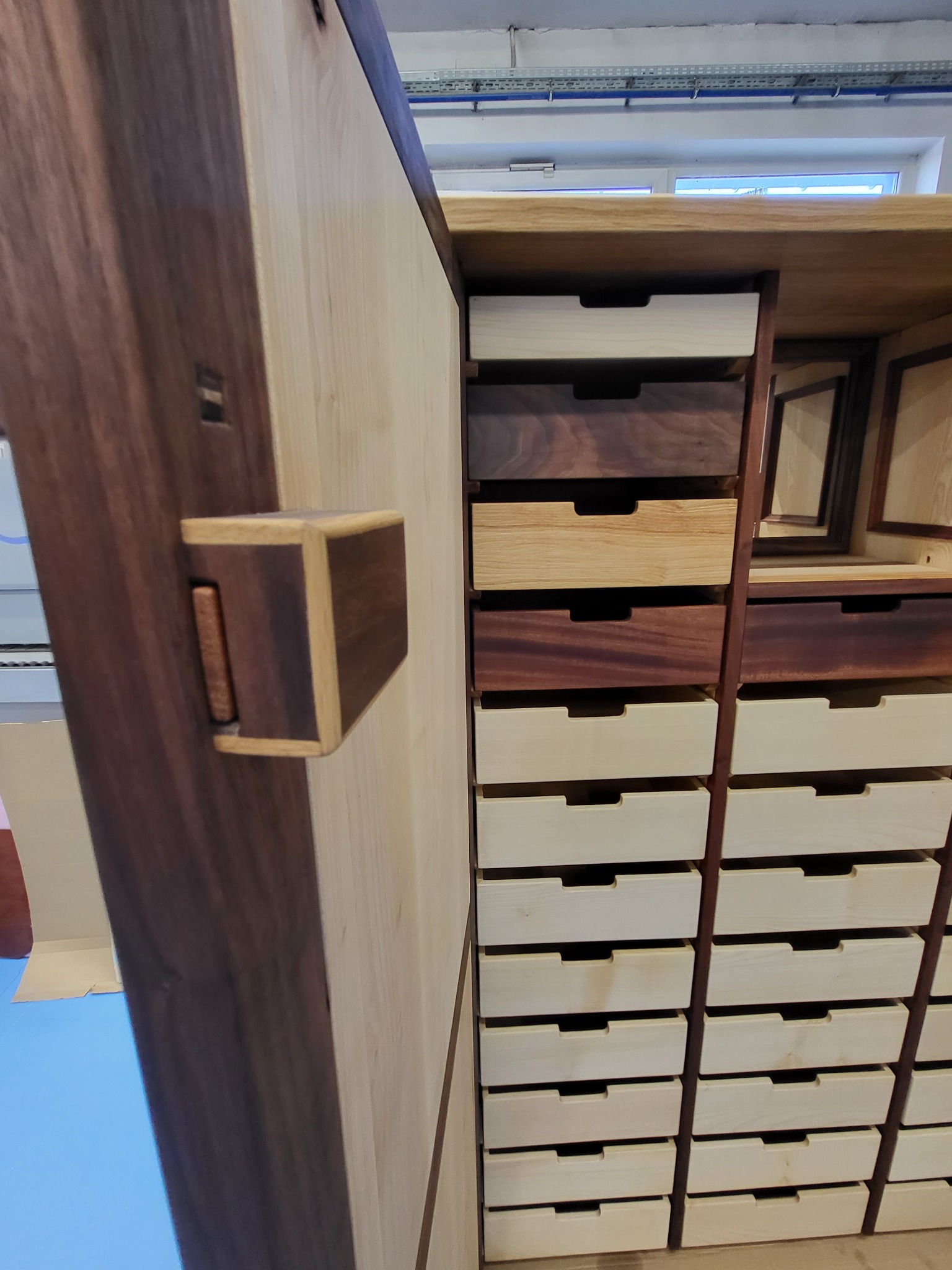 Drewniana szafa z otwartymi drzwiami, prezentująca różnorodne szuflady z jasnego i ciemnego drewna, z widocznym detalem zamka.