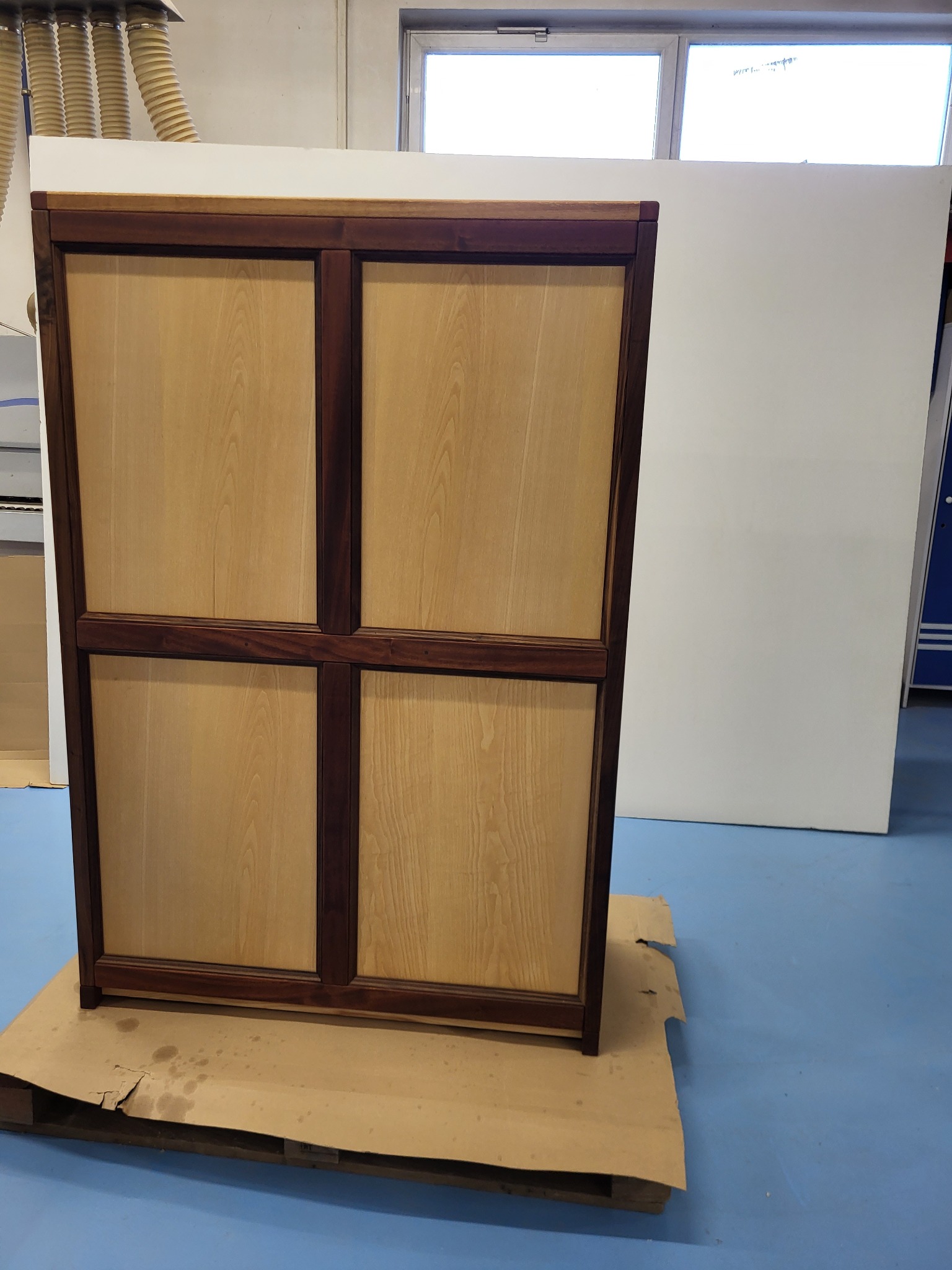 Drewniana szafka z czterema panelami o jasnym, naturalnym odcieniu, obramowana ciemnym drewnem, stoi na palecie w warsztacie. Widoczne rury wentylacyjne.