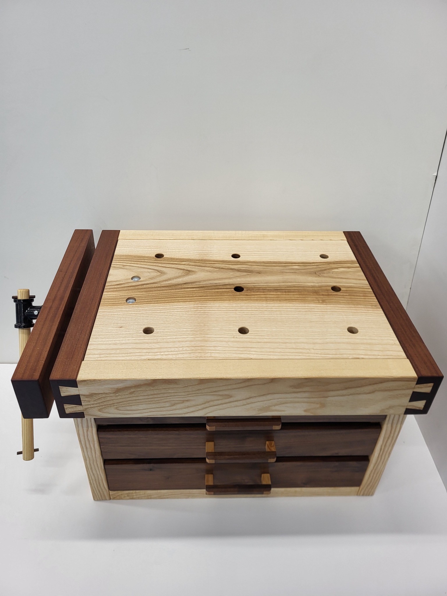 Drewniany stół warsztatowy z jasnego drewna z ciemnobrązowymi detalami, widoczne szuflady i imadło. Charakterystyczne połączenia elementów na rogach blatu.
