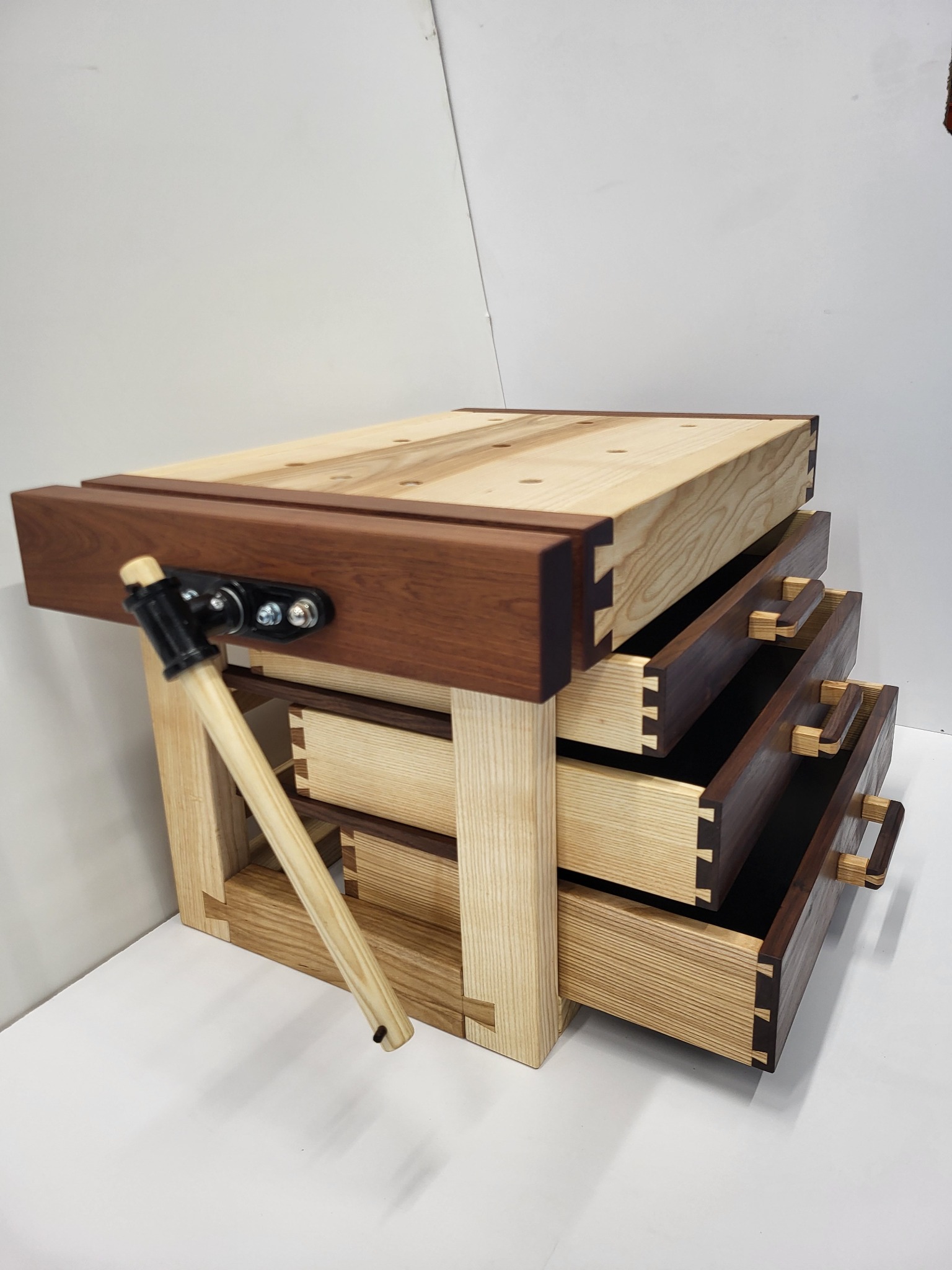 Drewniany stół warsztatowy z trzema szufladami, widoczne połączenia na jaskółczy ogon, z imadłem i młotkiem, wykonany z różnych gatunków drewna.