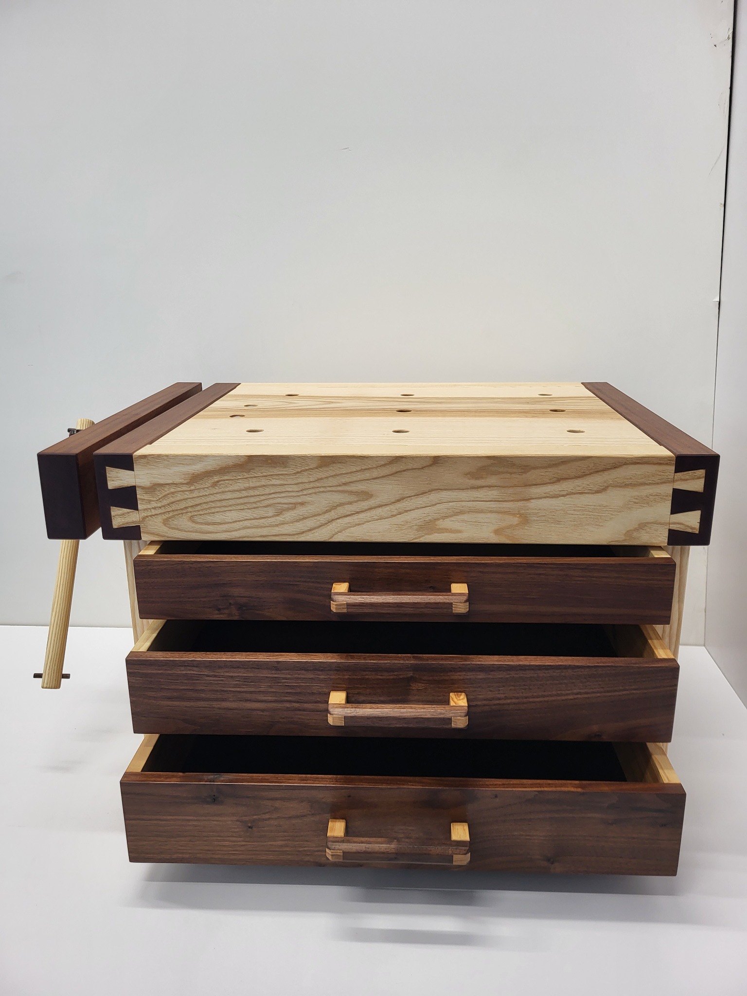 Drewniany stół warsztatowy z trzema szufladami i imadłem, wykonany z jasnego i ciemnego drewna, z widocznymi połączeniami stolarskimi typu jaskółczy ogon.