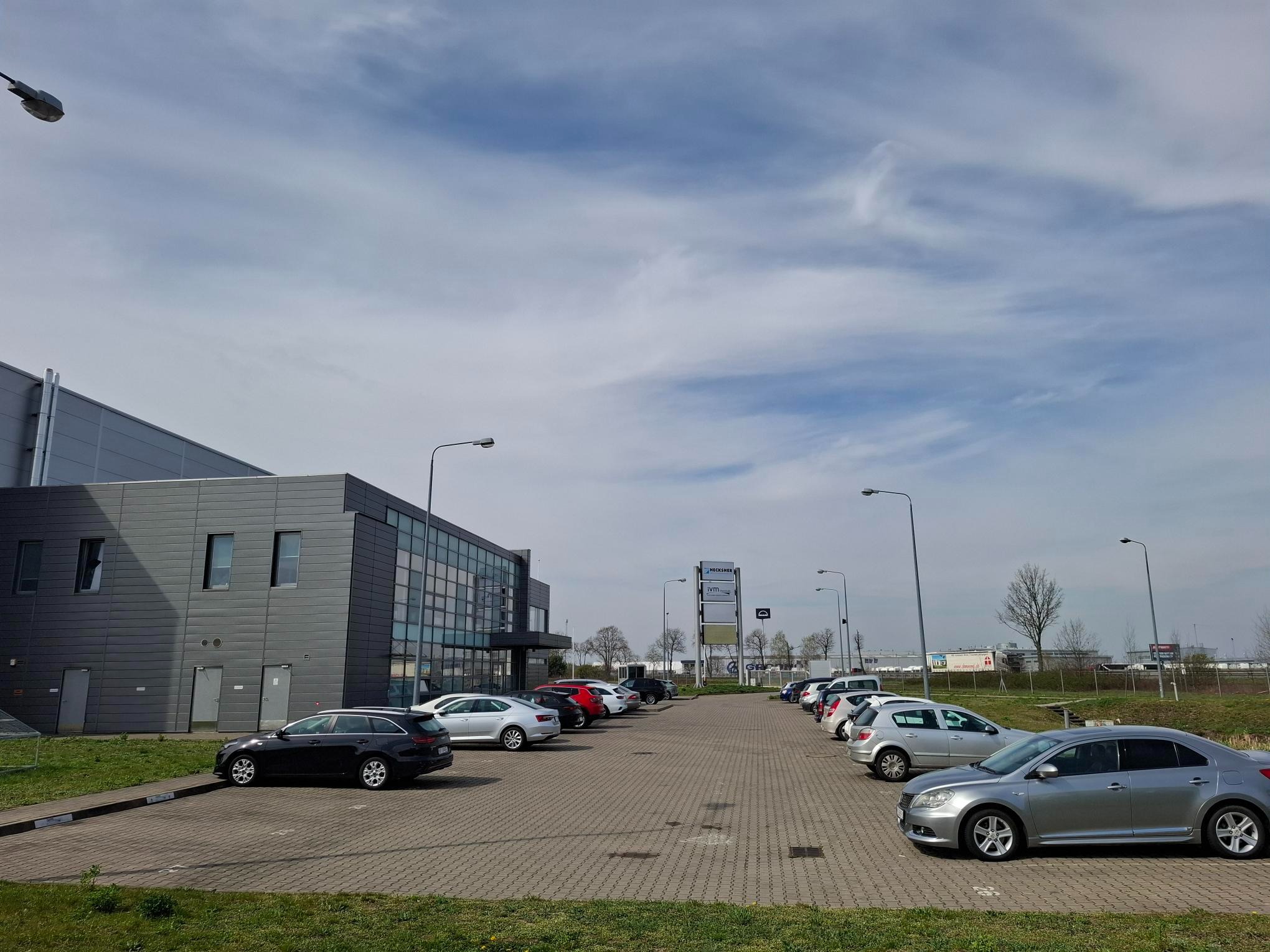 Szary budynek magazynowy z parkingiem pełnym samochodów w słoneczny dzień. Widoczny znak firmowy na tle błękitnego nieba z chmurami.