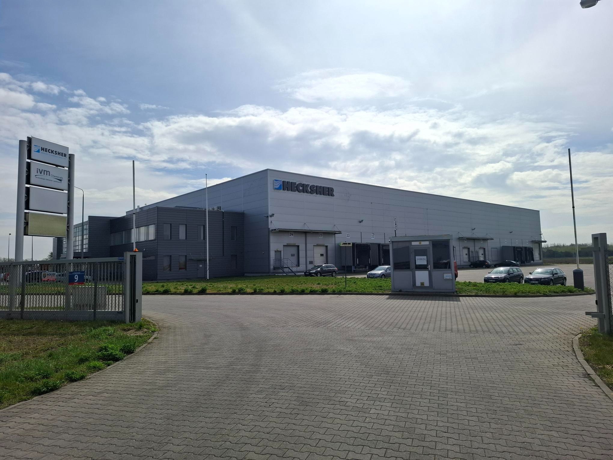 Szary budynek magazynowy Hecksher z rampami załadunkowymi, widoczny z zewnątrz, tablica informacyjna i szary bruk na pierwszym planie.