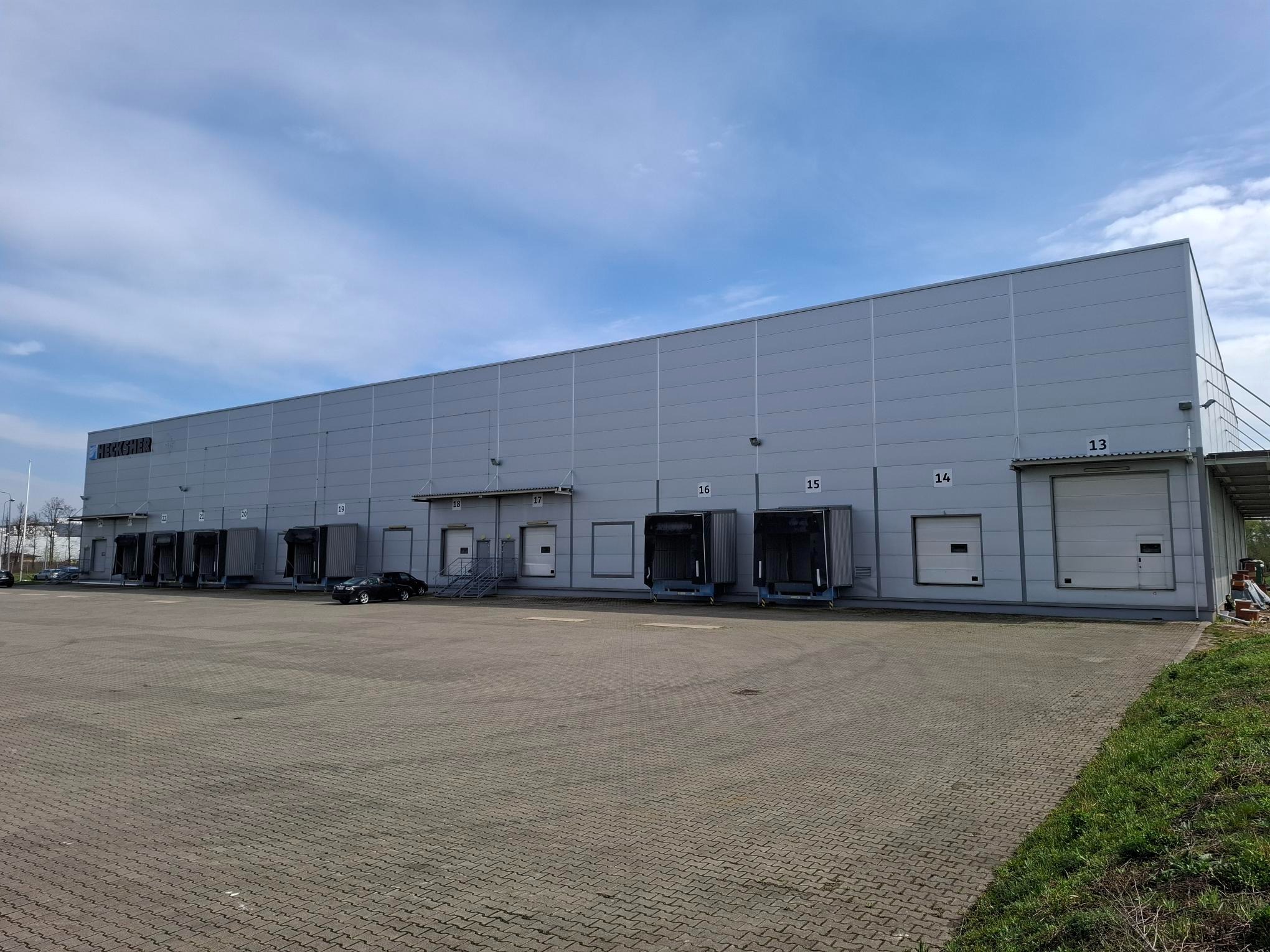 Duży, szary magazyn z rampami załadunkowymi o numerach 13-16. Na placu manewrowym zaparkowany samochód. Błękitne niebo z delikatnymi chmurami w tle.