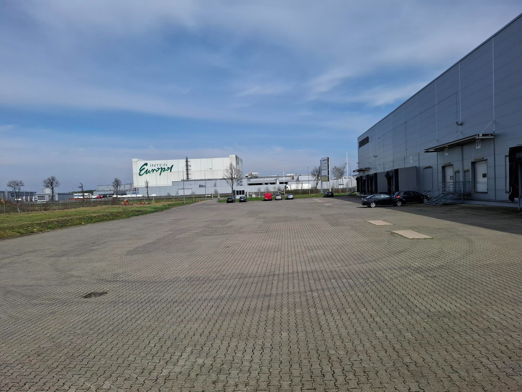 Rozległy magazyn Inter Europol z rampami załadunkowymi, widoczny na tle błękitnego nieba z lekkimi chmurami. Duży plac wyłożony kostką brukową na pierwszym planie.
