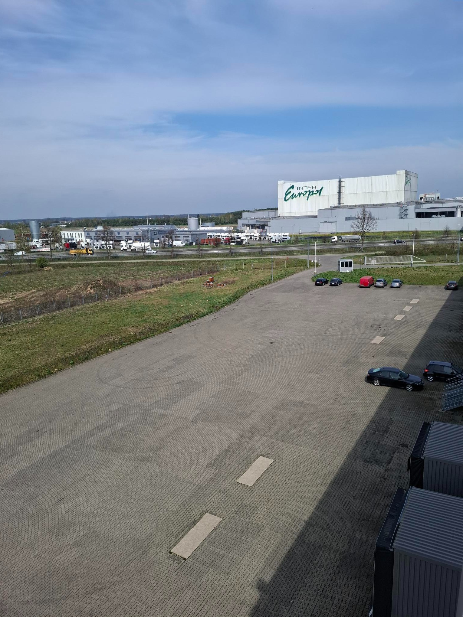 Szeroki kadr na budynek magazynowy Inter Europol z parkingiem, otoczony zielenią i innymi budynkami w tle, w słoneczny dzień.