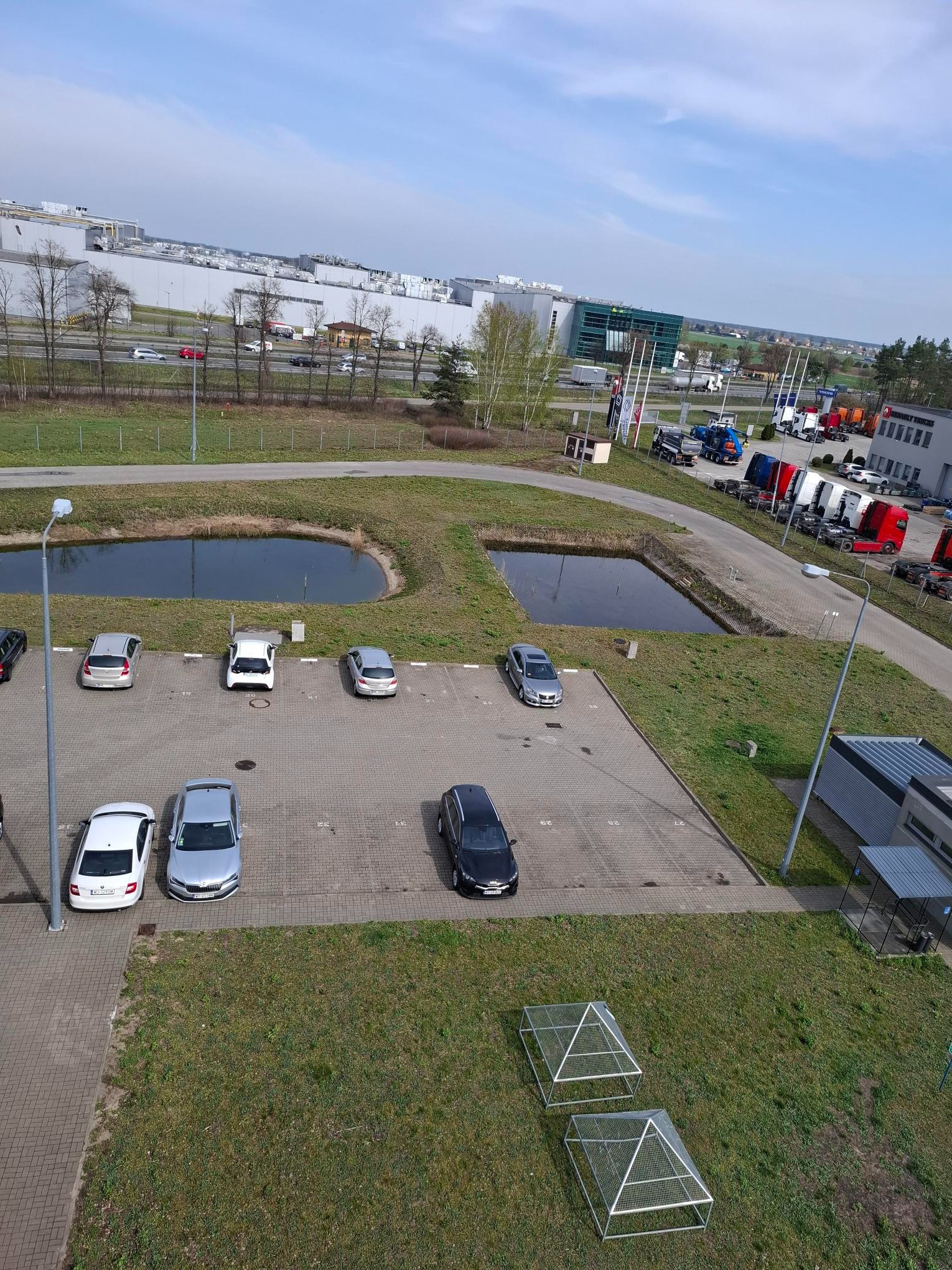 Widok z góry na parking samochodowy z budynkiem magazynowym w tle, dwa stawy i konstrukcje szkieletowe na trawie. Dzień słoneczny, błękitne niebo.
