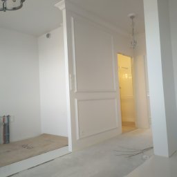 Glazrem - Jasne wnętrze w trakcie wykańczania: białe ściany z dekoracyjnymi listwami, surowe podłogi, widoczne instalacje. W tle oświetlone pomieszczenie.