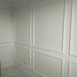 Glazrem - Eleganckie, białe panele ścienne z dekoracyjnymi listwami tworzącymi geometryczne wzory. Widoczny fragment podłogi i narożnik pomieszczenia. Klasyczny design.