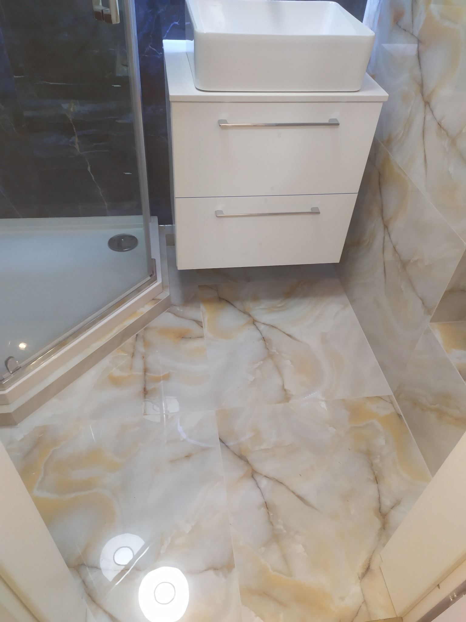 Wykończona łazienka z kabiną prysznicową, białą szafką pod umywalką i marmurową okładziną ścian oraz podłogi w odcieniach beżu i brązu. Nowoczesny design.