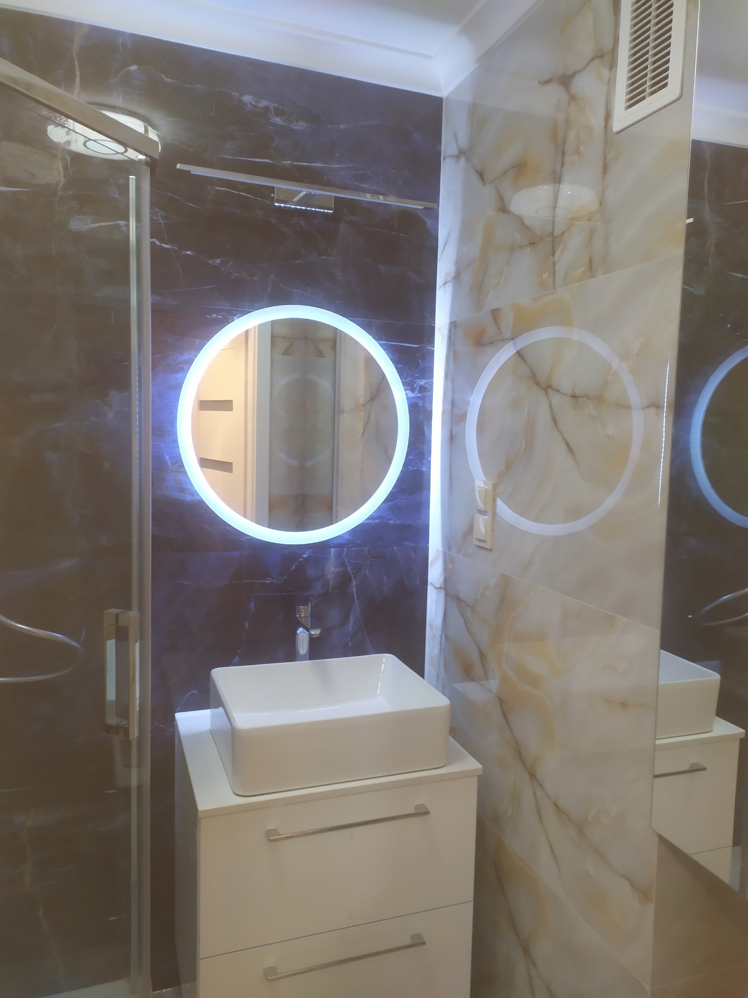 Nowoczesna łazienka z umywalką nablatową, podświetlanym lustrem i marmurowymi płytkami. Kabina prysznicowa ze szklanymi drzwiami po lewej stronie.