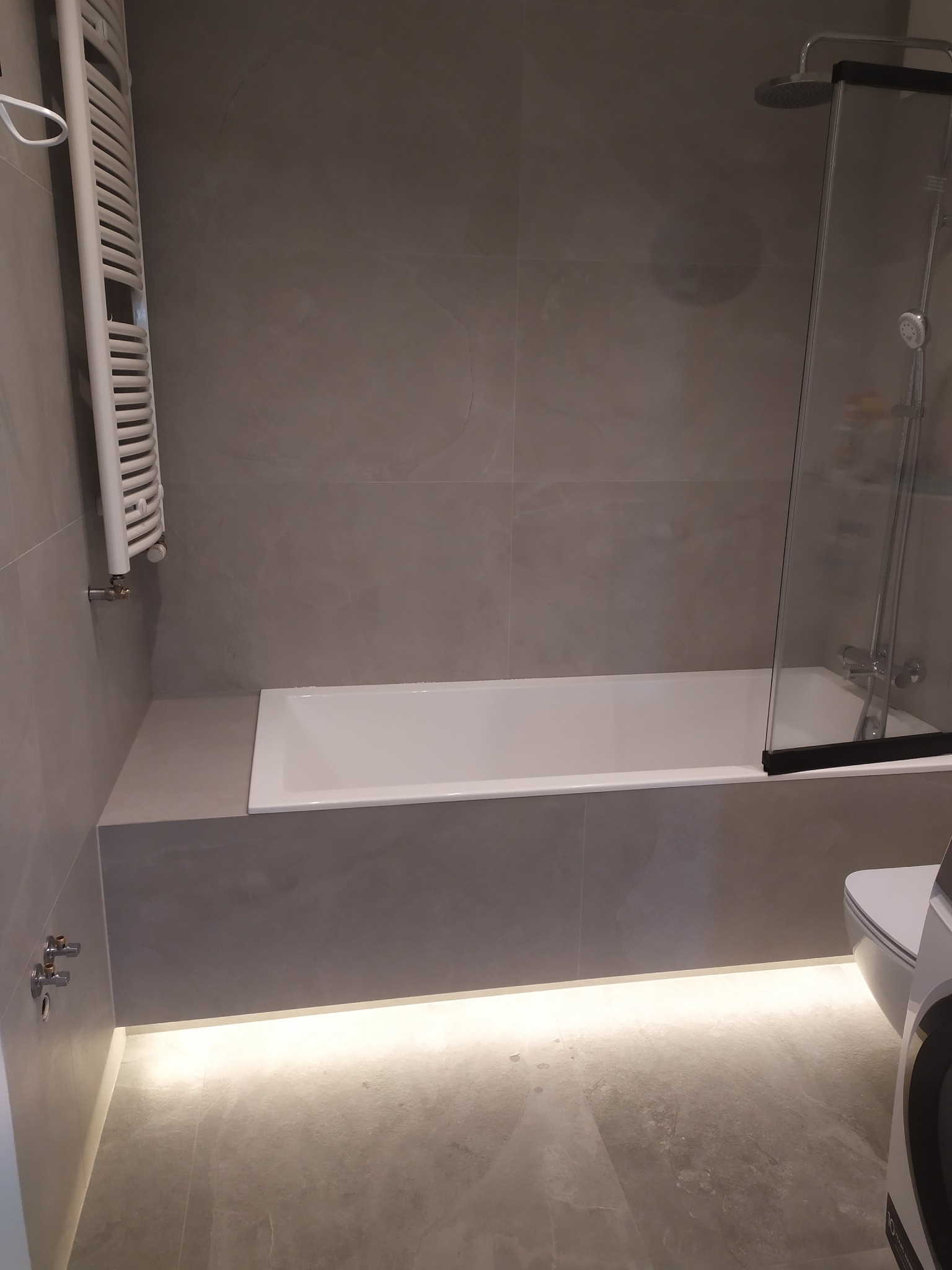 Nowoczesna łazienka w szarościach z podświetlaną wanną, grzejnikiem drabinkowym i kabiną prysznicową. Minimalistyczny design z dbałością o detale.