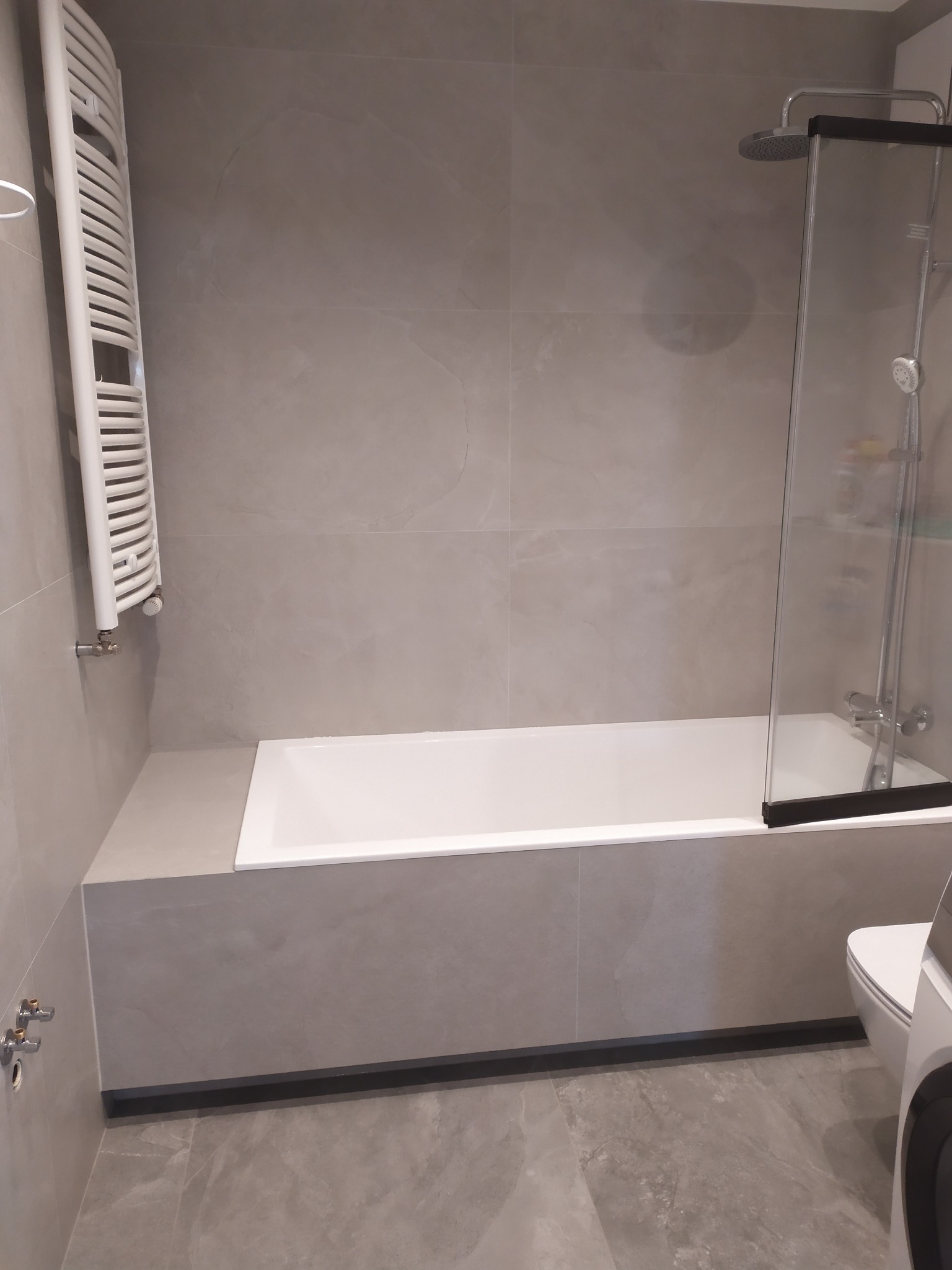 Nowoczesna łazienka w szarościach: wanna z parawanem, grzejnik drabinkowy i płytki imitujące kamień. Minimalistyczny design i funkcjonalne rozwiązania.