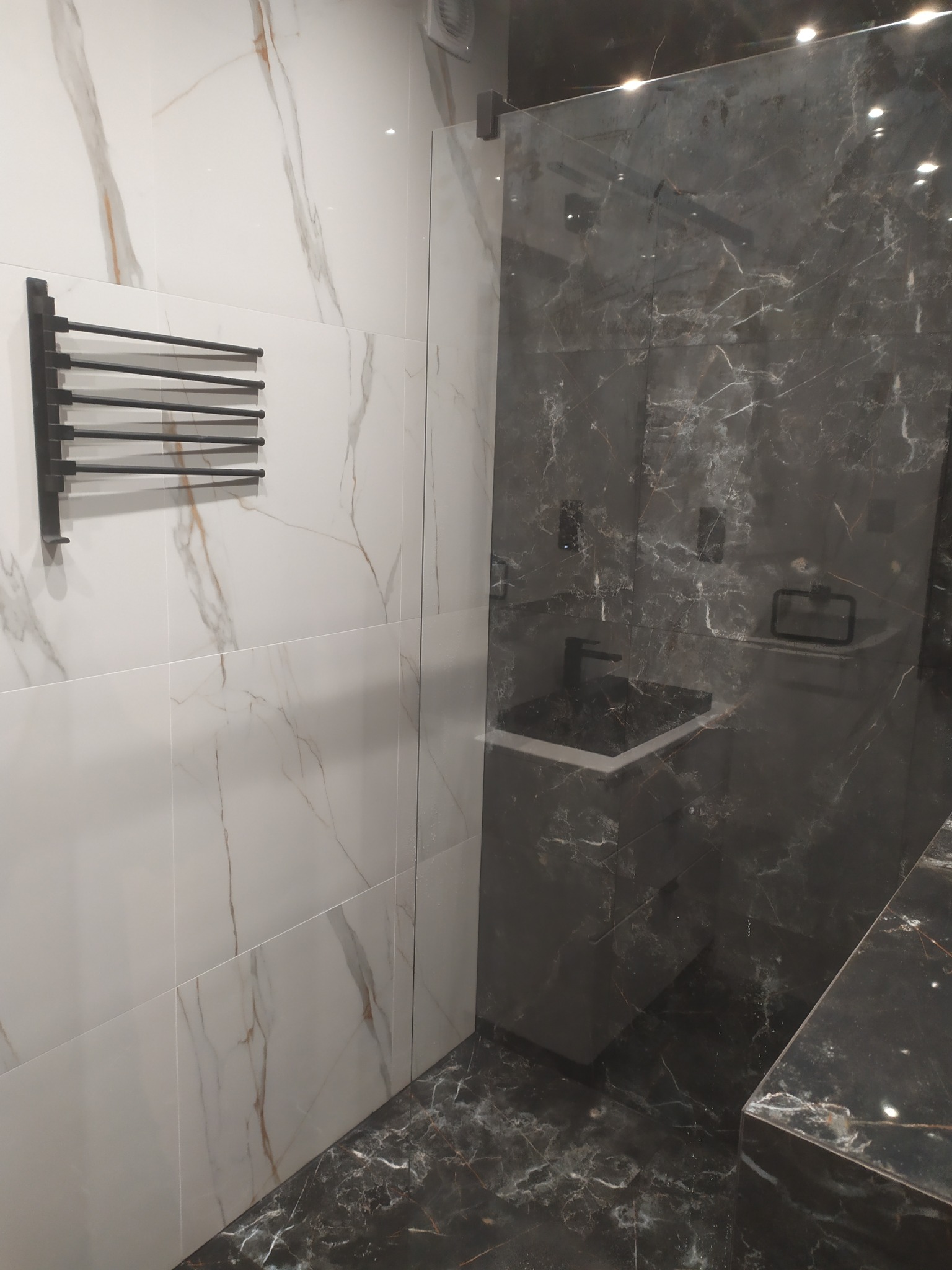 Nowoczesna łazienka z marmurowymi płytkami w odcieniach bieli i szarości. Czarny grzejnik drabinkowy i szklana kabina prysznicowa dodają elegancji.