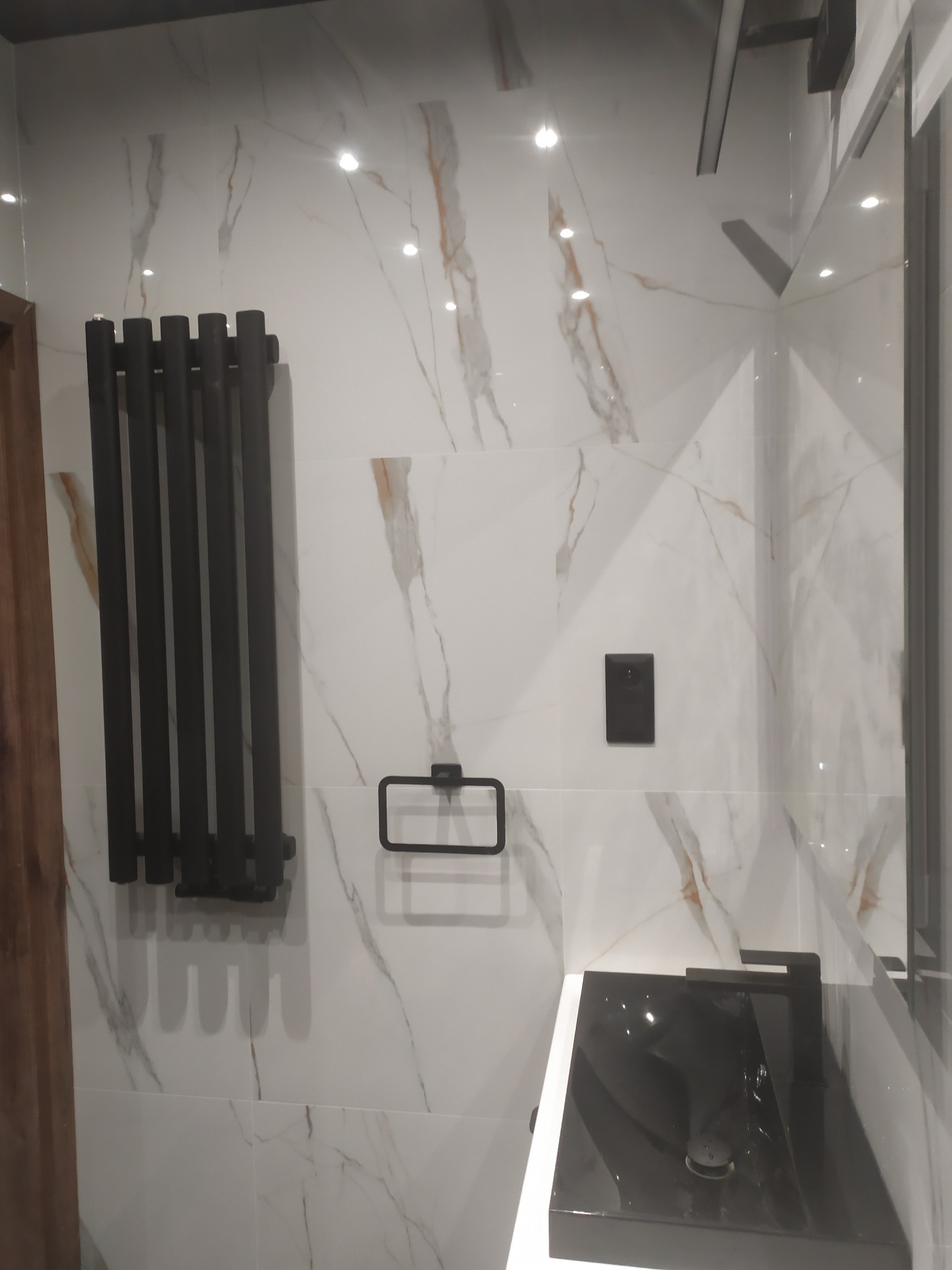 Nowoczesna łazienka z czarną armaturą: grzejnik, bateria umywalkowa i uchwyt na ręcznik na tle białych płytek imitujących marmur z delikatnymi żyłkami.