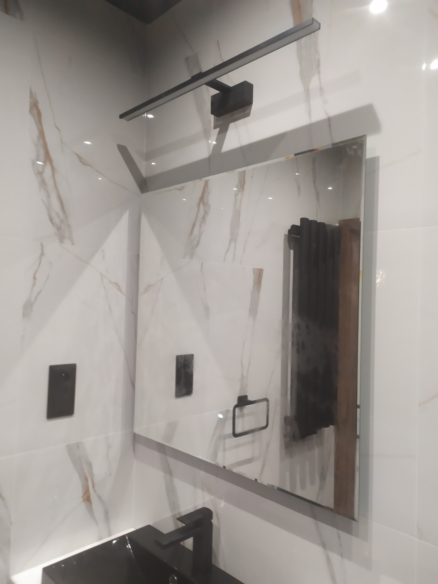 Wykończona łazienka z marmurowymi płytkami, czarną armaturą i oświetleniem LED nad lustrem. Widoczne detale: włączniki i uchwyt na ręcznik. Nowoczesny design.