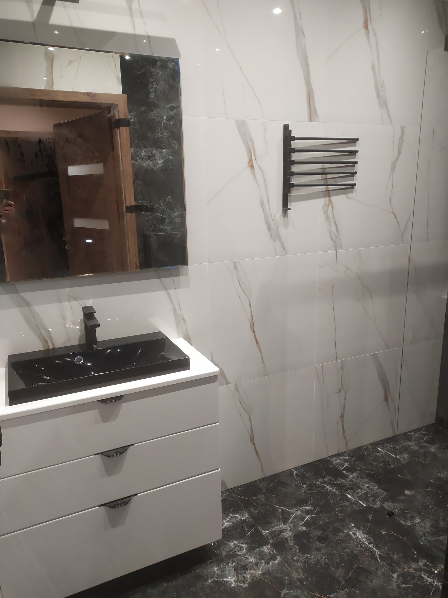 Nowoczesna łazienka z czarną umywalką na białej szafce, marmurowe płytki ścienne i podłogowe, oraz czarny wieszak na ręczniki. Lustro z drewnianą ramą.