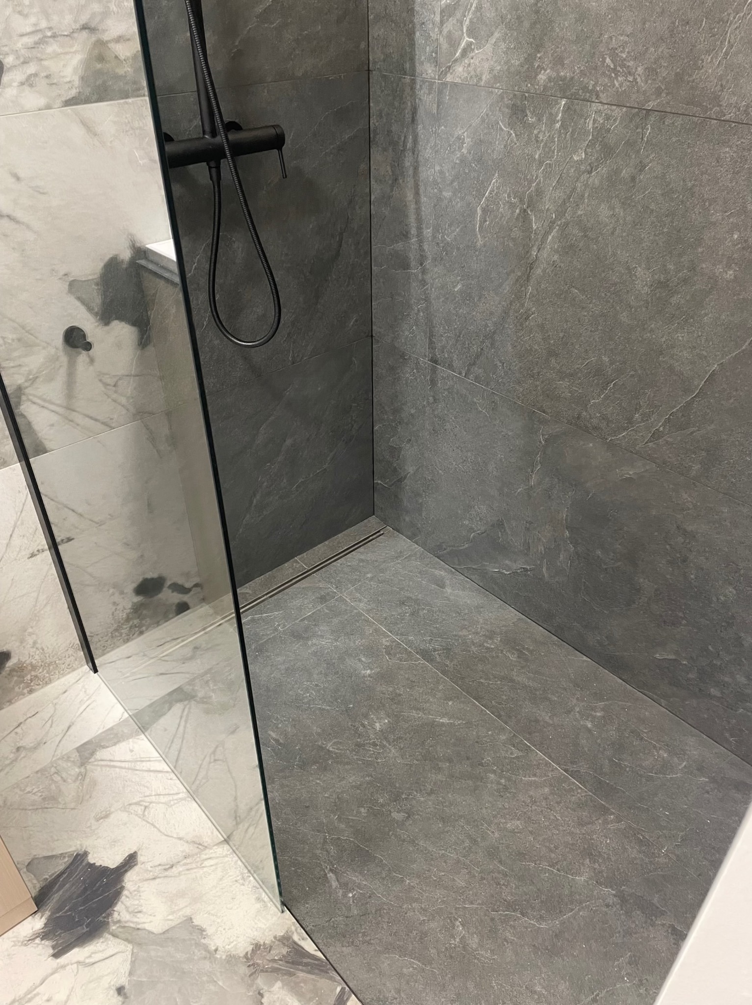 Nowoczesna kabina prysznicowa z szarymi płytkami imitującymi kamień i czarną armaturą. Widoczny odpływ liniowy i fragment szklanej ścianki.