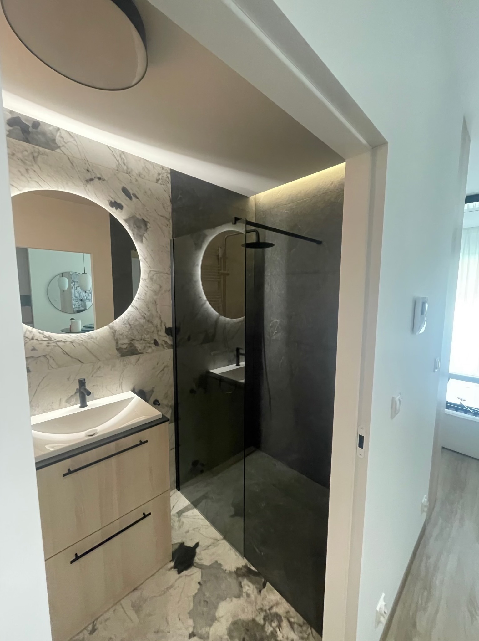 Nowoczesna łazienka z marmurowymi płytkami, okrągłym lustrem z podświetleniem LED i kabiną prysznicową typu walk-in z czarną armaturą.