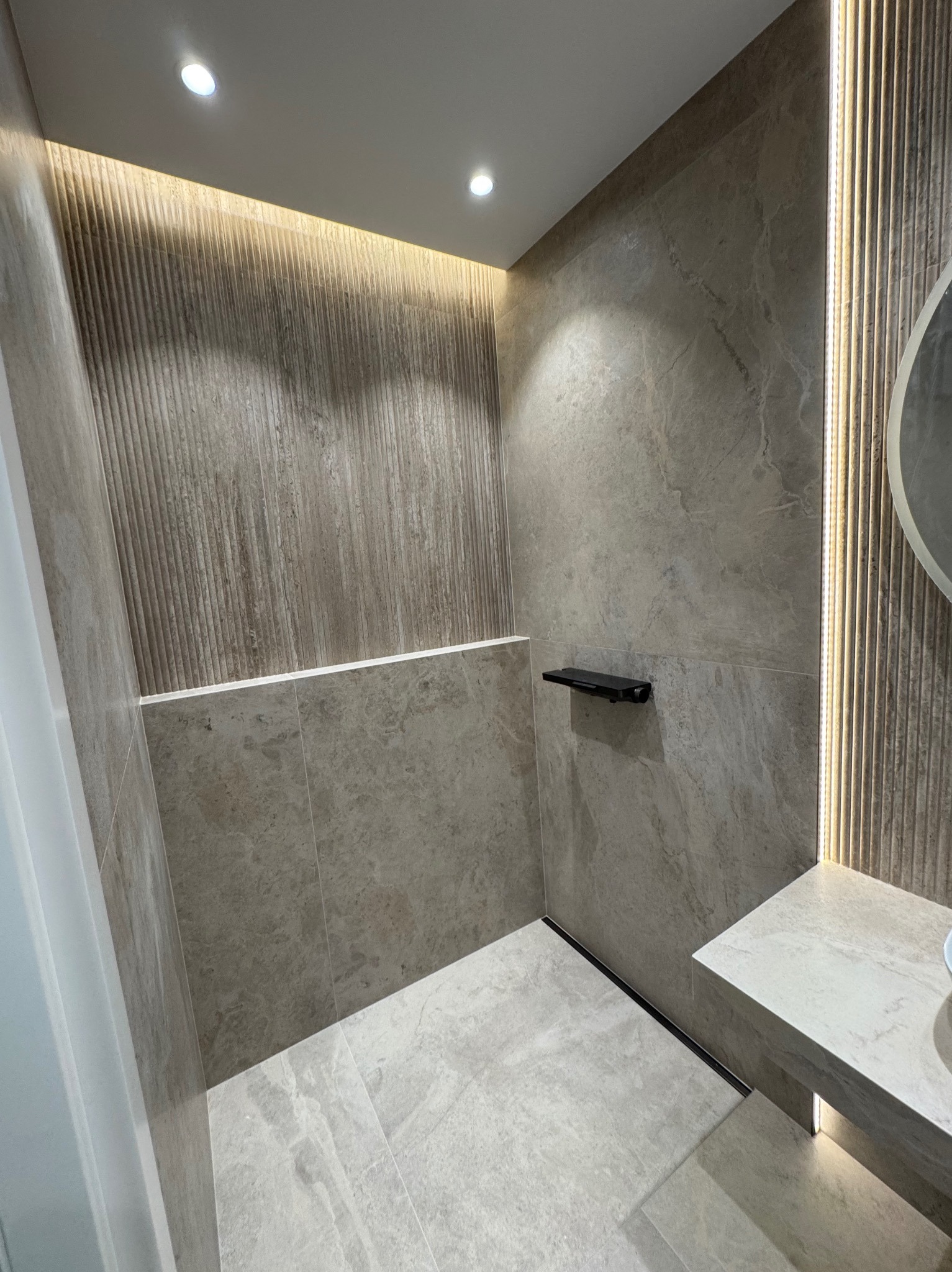 Wykończona łazienka z beżowymi płytkami imitującymi kamień, oświetleniem LED i czarnymi akcentami. Widoczna półka i fragment umywalki. Minimalistyczny design.