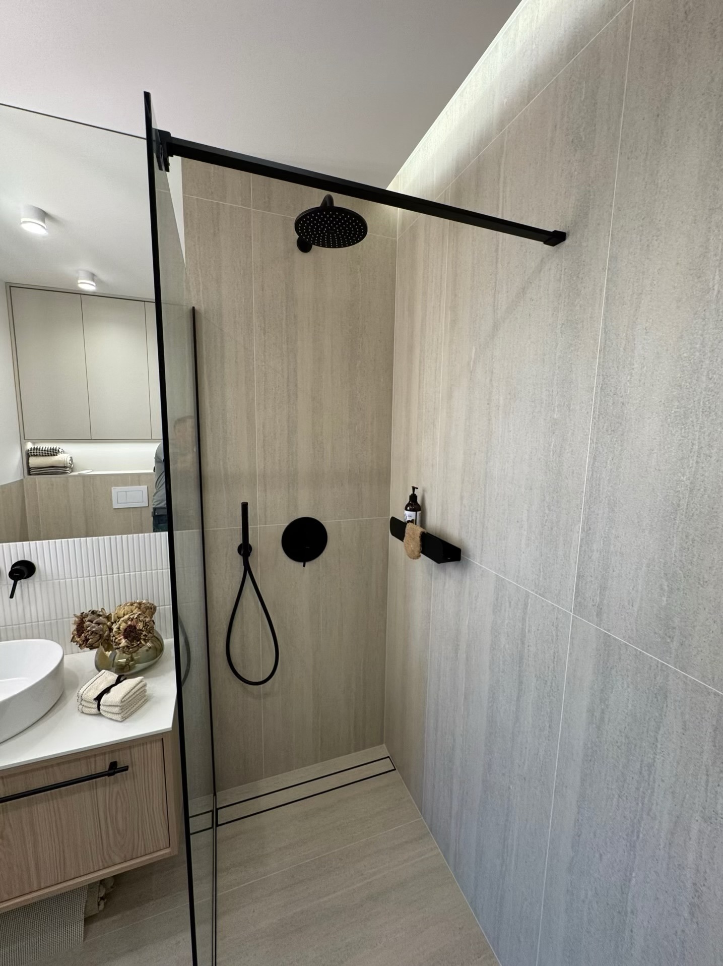 Nowoczesna łazienka z kabiną typu walk-in, wykończona płytkami w odcieniach szarości. Czarna armatura, minimalistyczny design, oświetlenie LED. Widoczny fragment umywalki i szafki.