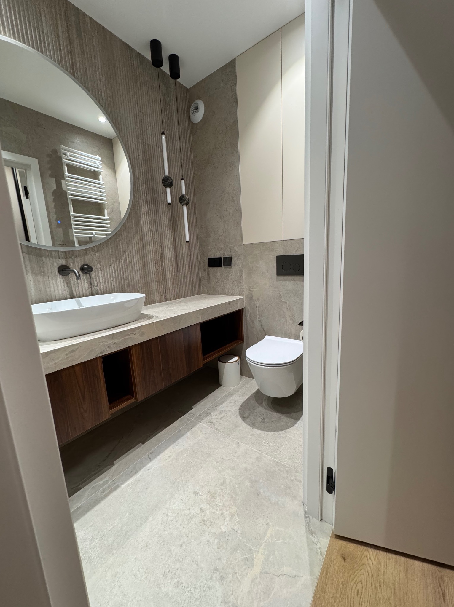 Nowoczesna łazienka z owalną umywalką nablatową, drewnianą szafką pod umywalką, wiszącą toaletą i szarą płytką imitującą kamień na ścianach i podłodze.
