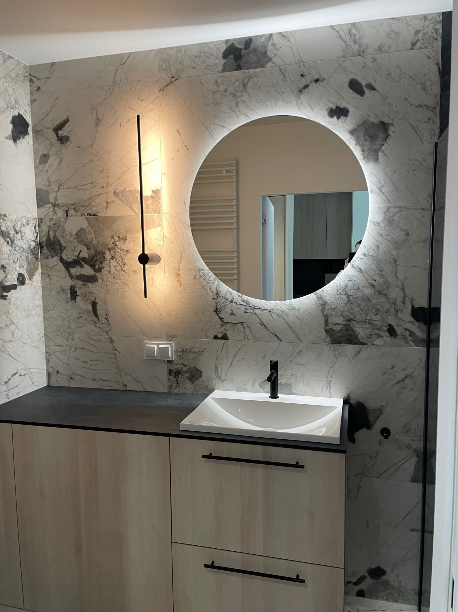 Nowoczesna łazienka z okrągłym lustrem LED i marmurowymi płytkami. Minimalistyczna szafka pod umywalką z czarnymi uchwytami. Oświetlenie punktowe i grzejnik w tle.